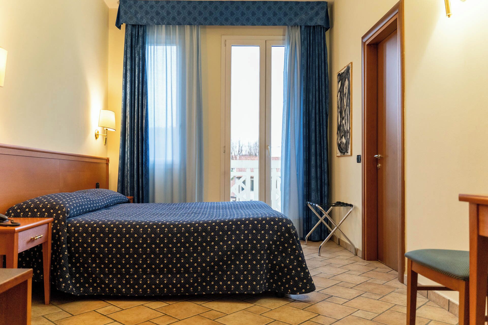 Una habitación de hotel con una cama, un escritorio, una silla, una lámpara y una puerta.