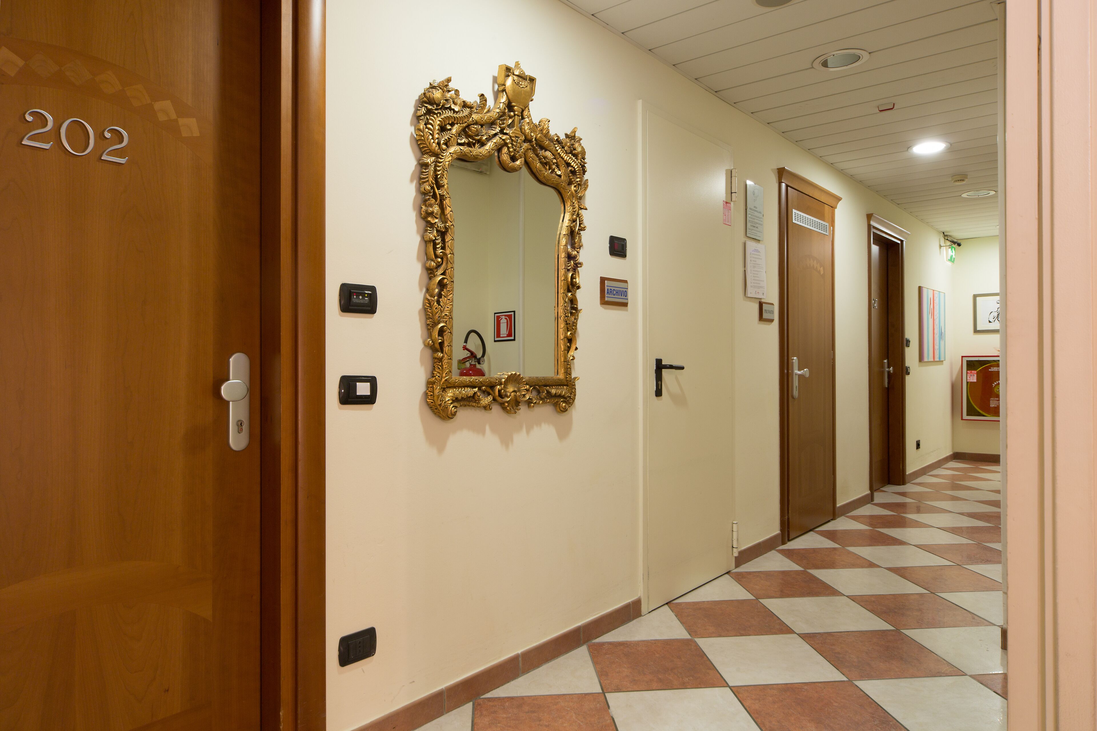 Un pasillo con piso a cuadros, un espejo grande y ornamentado, y puertas numeradas 202.