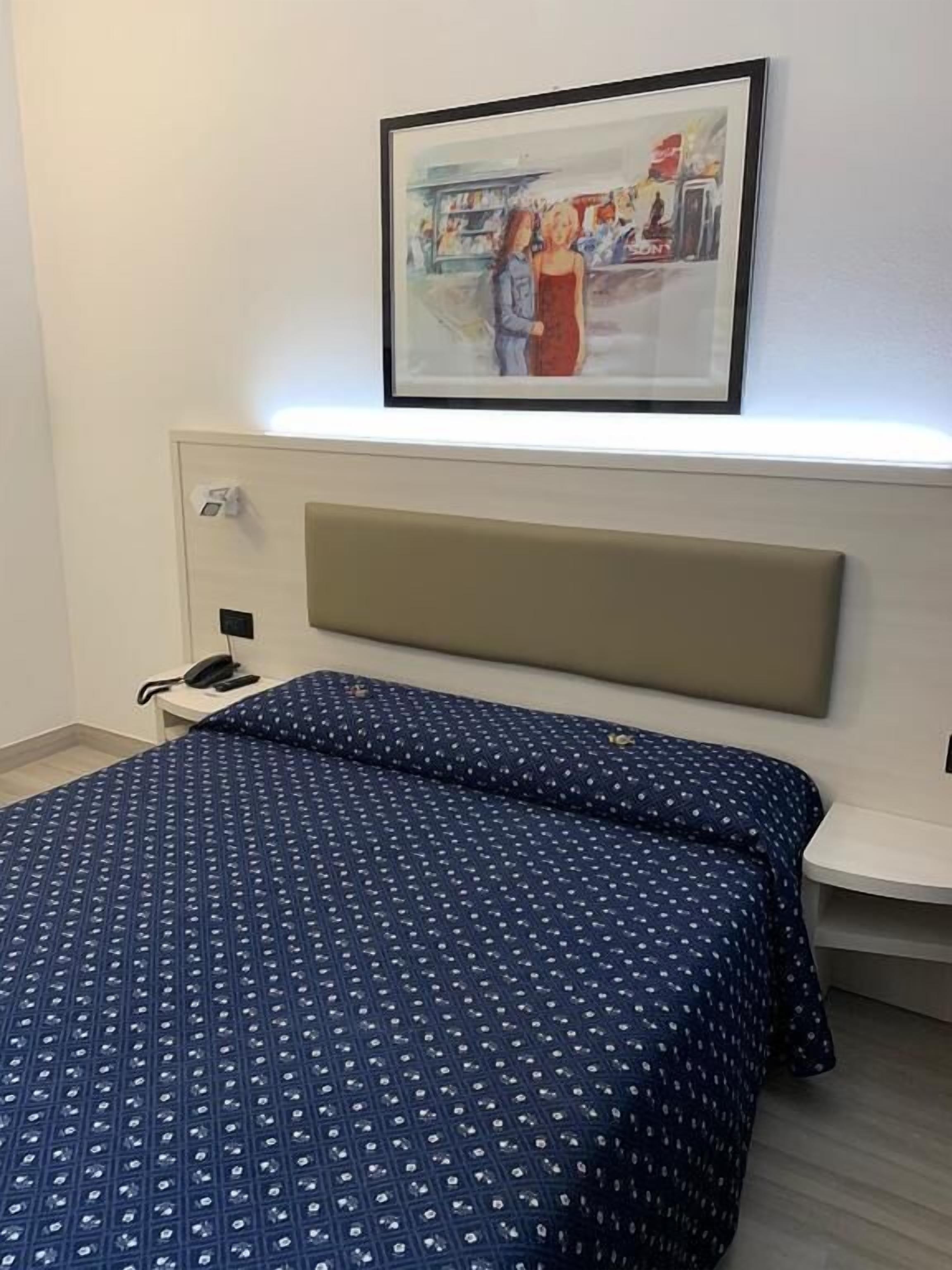 Een hotelkamer met een bed, nachtkastje, telefoon en een ingelijst schilderij aan de muur.
