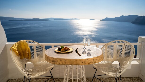 A balcony with a round table, two white wicker chairs, a plate of fruit, and two glasses of water. - Delfini Villas (Santorini)