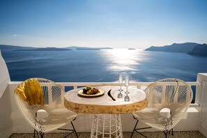 A round table with a plate of fruit and two glasses, two white wicker chairs, and a view of the sea. - Delfini Villas (Santorini)