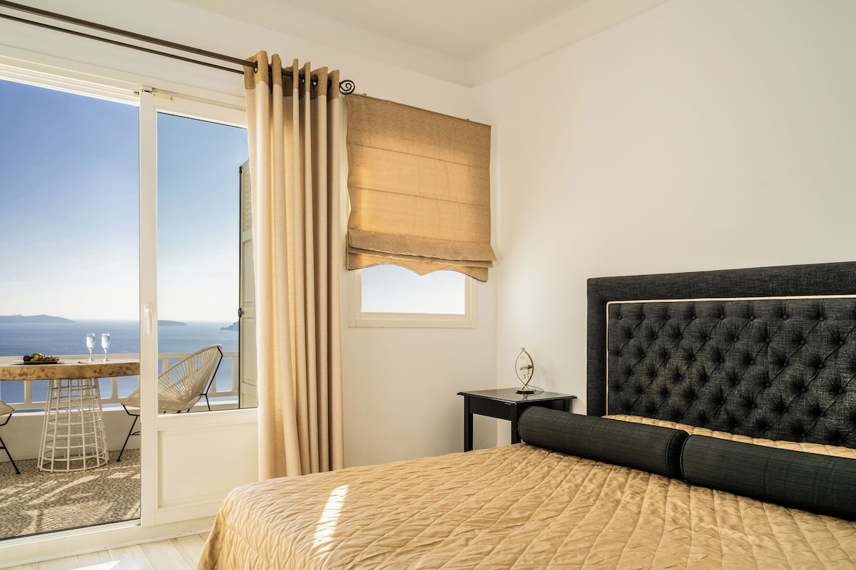 Ein Schlafzimmer mit einem großen Bett, einem Nachttisch, einem Fenster mit Vorhängen, Meerblick und einem Balkon mit Tisch und Stühlen.