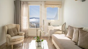 A room with a sofa, armchair, and a small table with a vase of flowers. There is a window with a view of the sea. - Delfini Villas (Santorini)