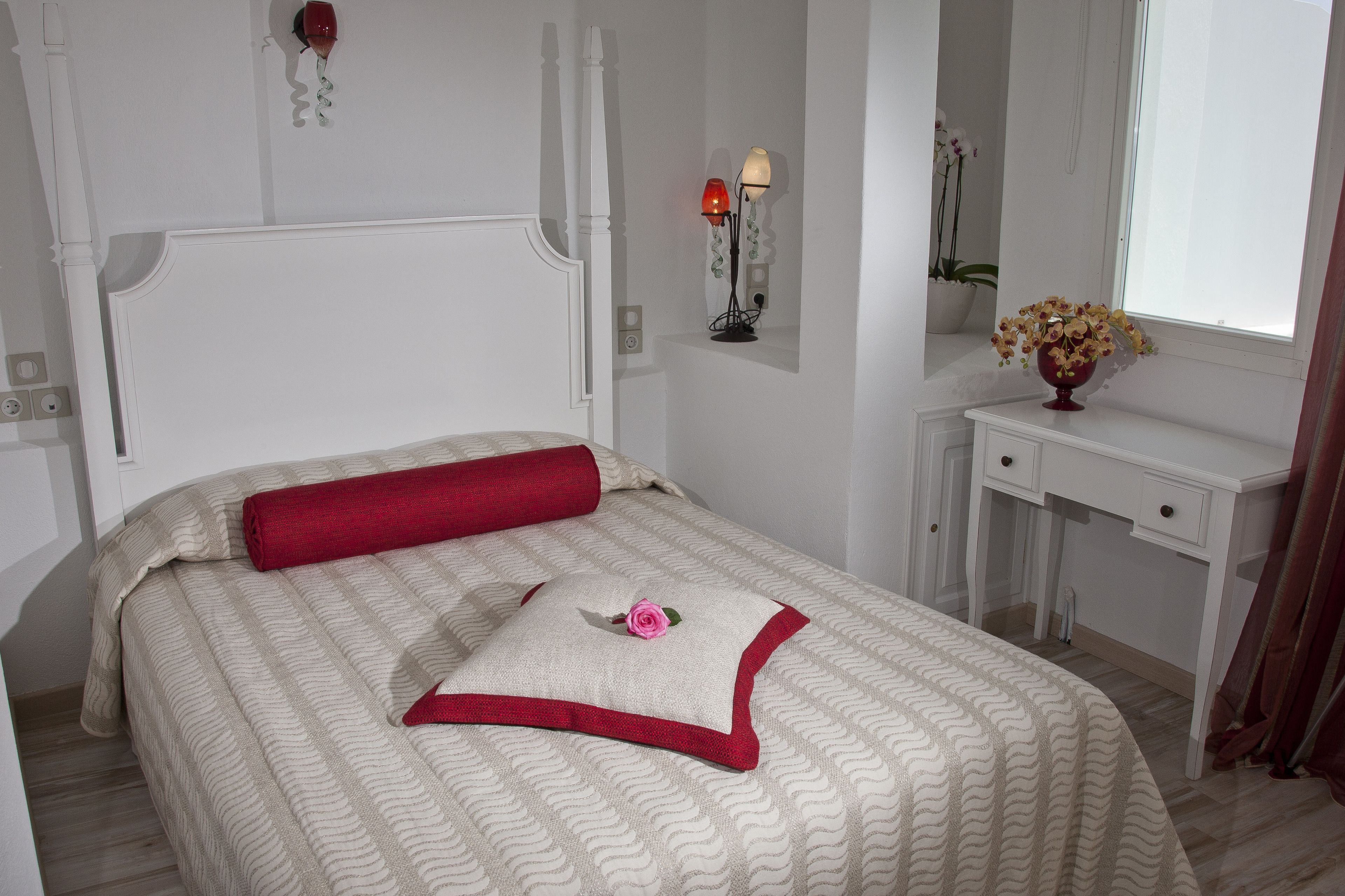 A neatly made bed with a red pillow and a decorative pillow with a flower. There is a bedside table with a lamp and a vase of flowers.