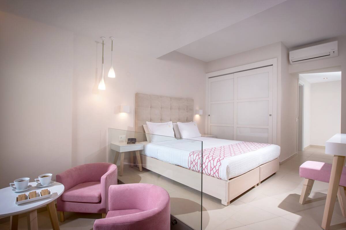 Een moderne hotelkamer met een bed, een bureau, twee roze stoelen en een klein tafeltje met kopjes en koekjes.