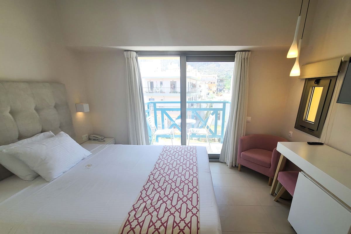 Een hotelkamer met een bed, een bureau, een stoel, een balkon met uitzicht op gebouwen en een raam met gordijnen.