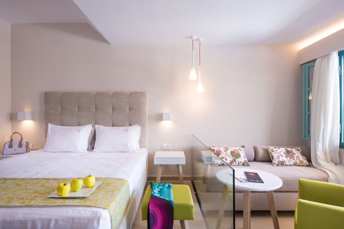 Een hotelkamer met een bed, nachtkastje, een kleintje eettafel met stoel, een bank en een raam met gordijnen.