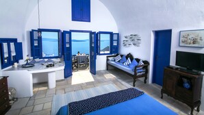 Una habitació amb un llit, un escriptori i una televisió. Hi ha finestres amb persianes blaves, una paret de pedra i vista al mar.