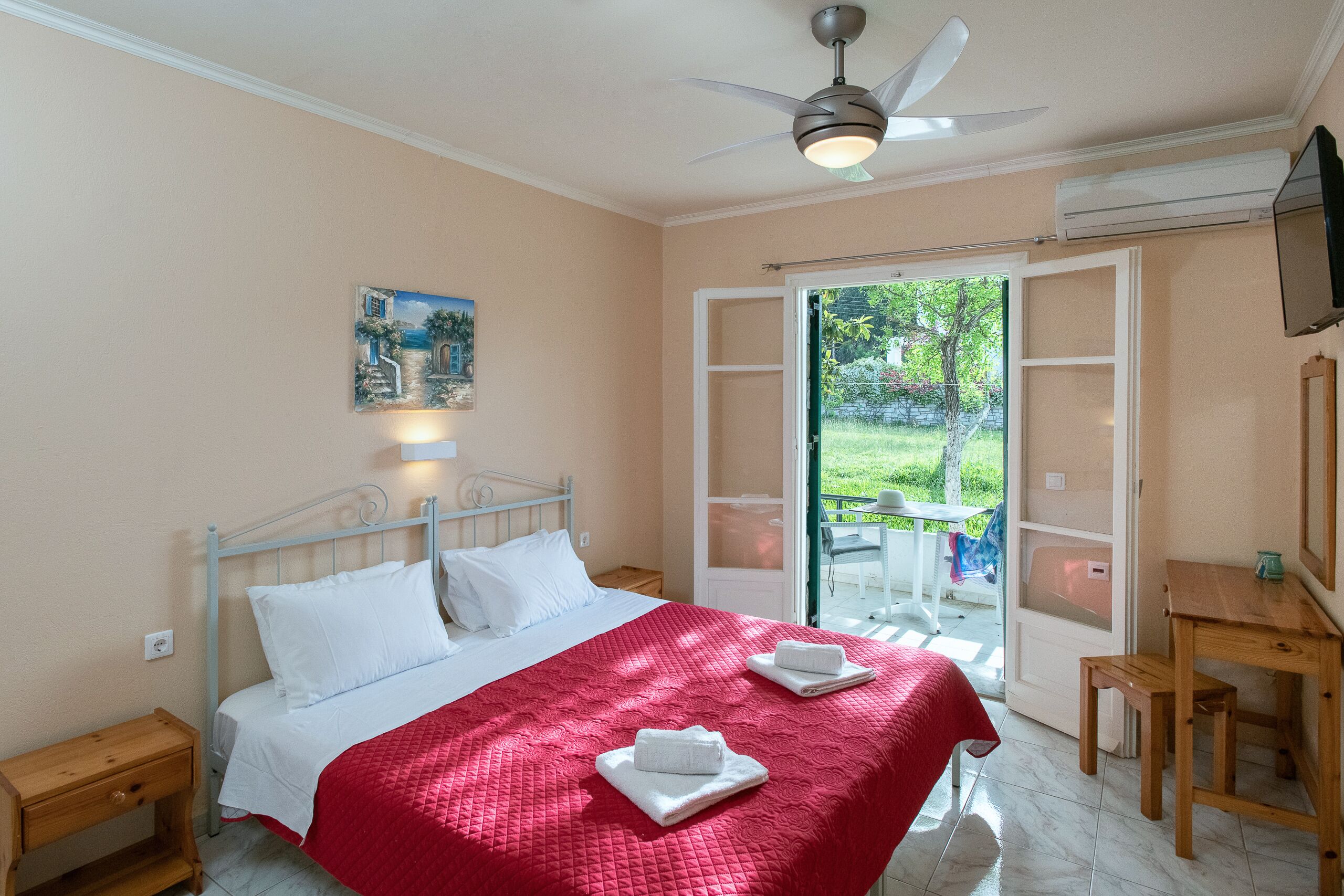A bedroom with a bed, a ceiling fan, a painting, a desk, and a view of the outdoors.