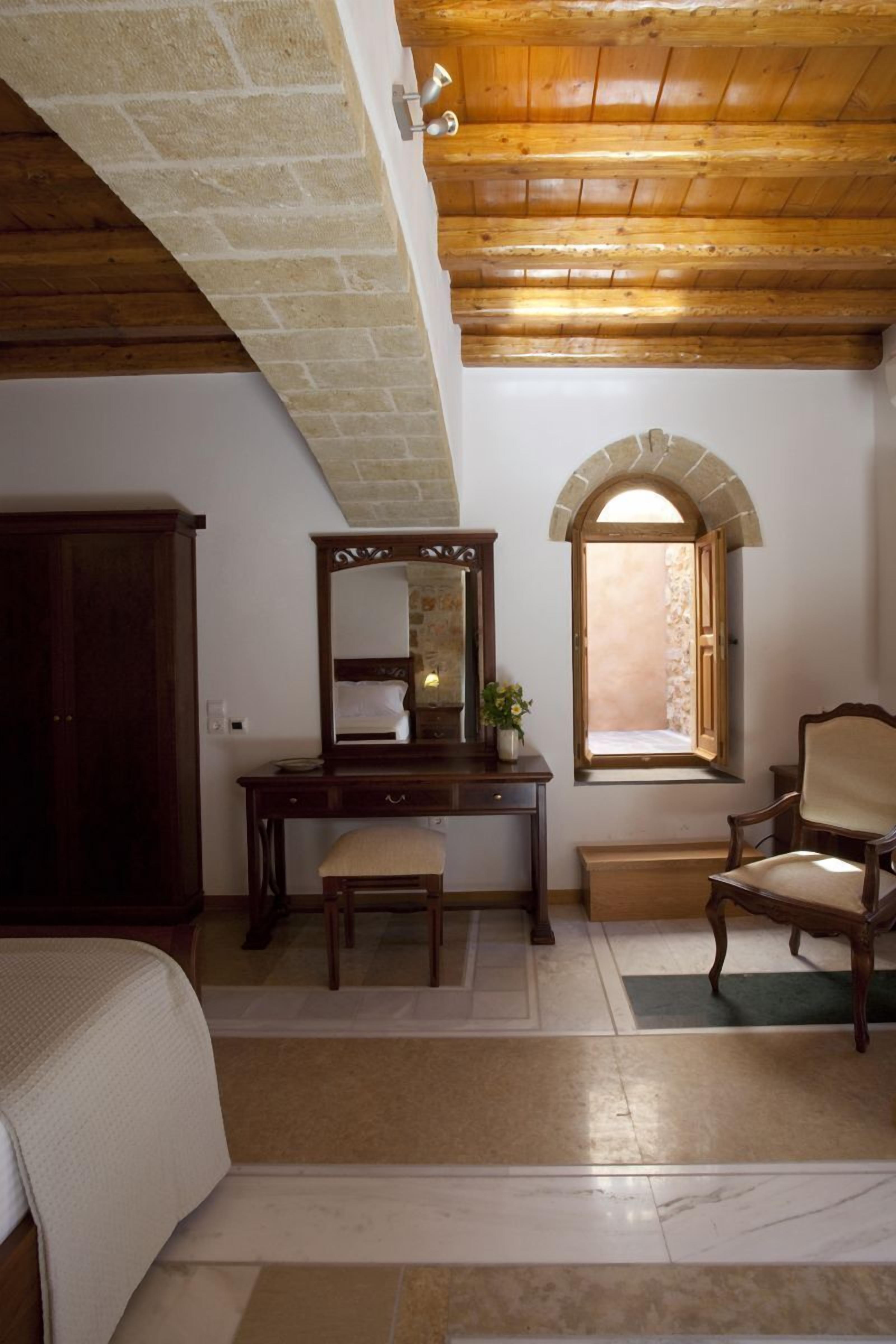 A room with a stone wall, wooden ceiling, a desk with a mirror, a chair, a bed, and a window with an arched top.