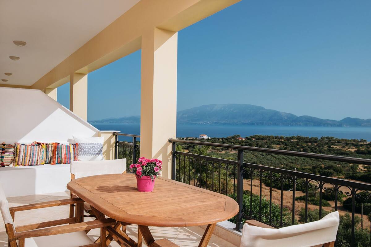 A balcony with a wooden table and chairs, a pink flower pot, and a view of the sea and mountains.