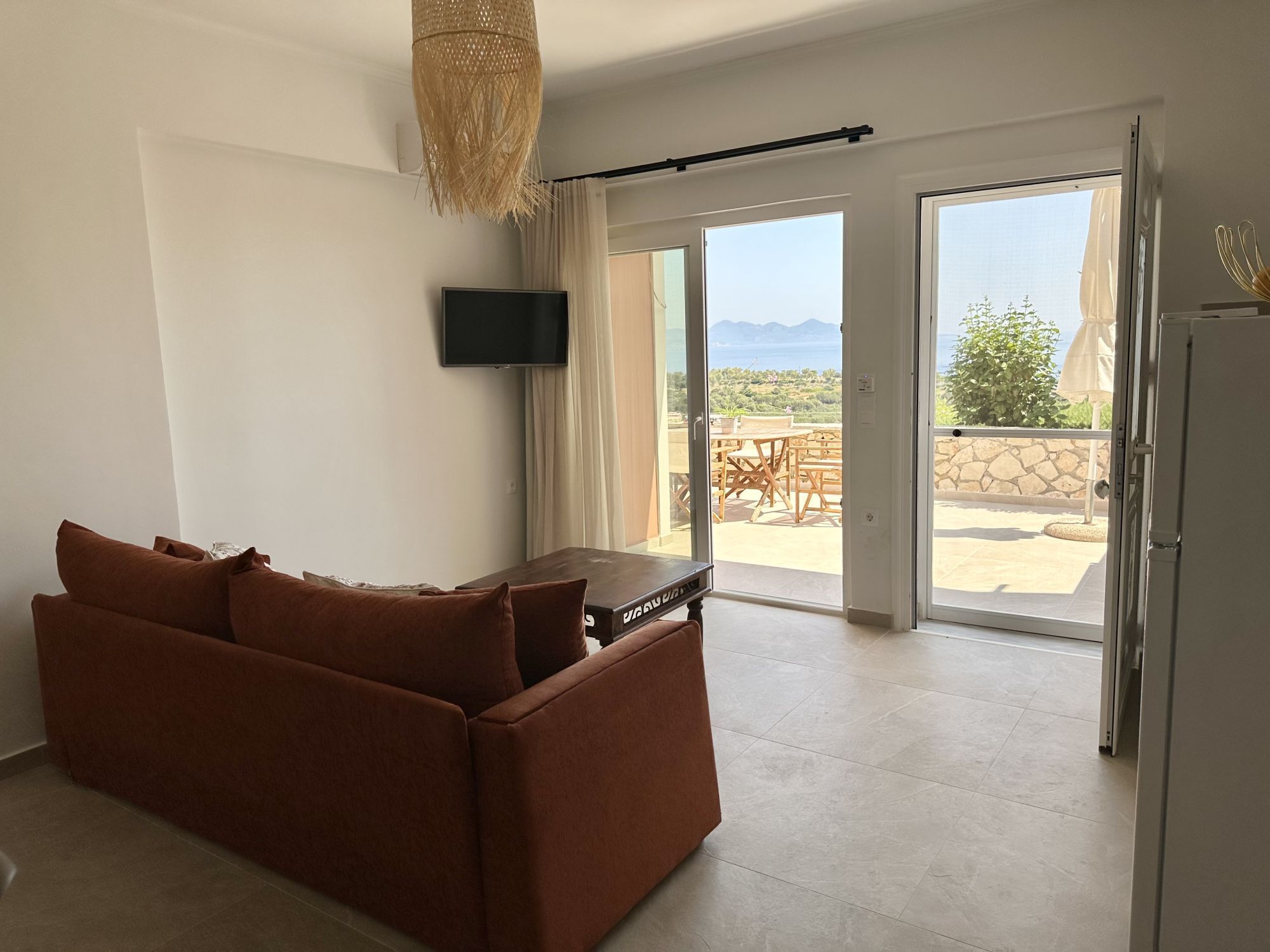 Ein Zimmer mit einem braunen Sofa, einem Flachbildfernseher und Blick auf eine Terrassengartenanlage mit Tisch und Stühlen.