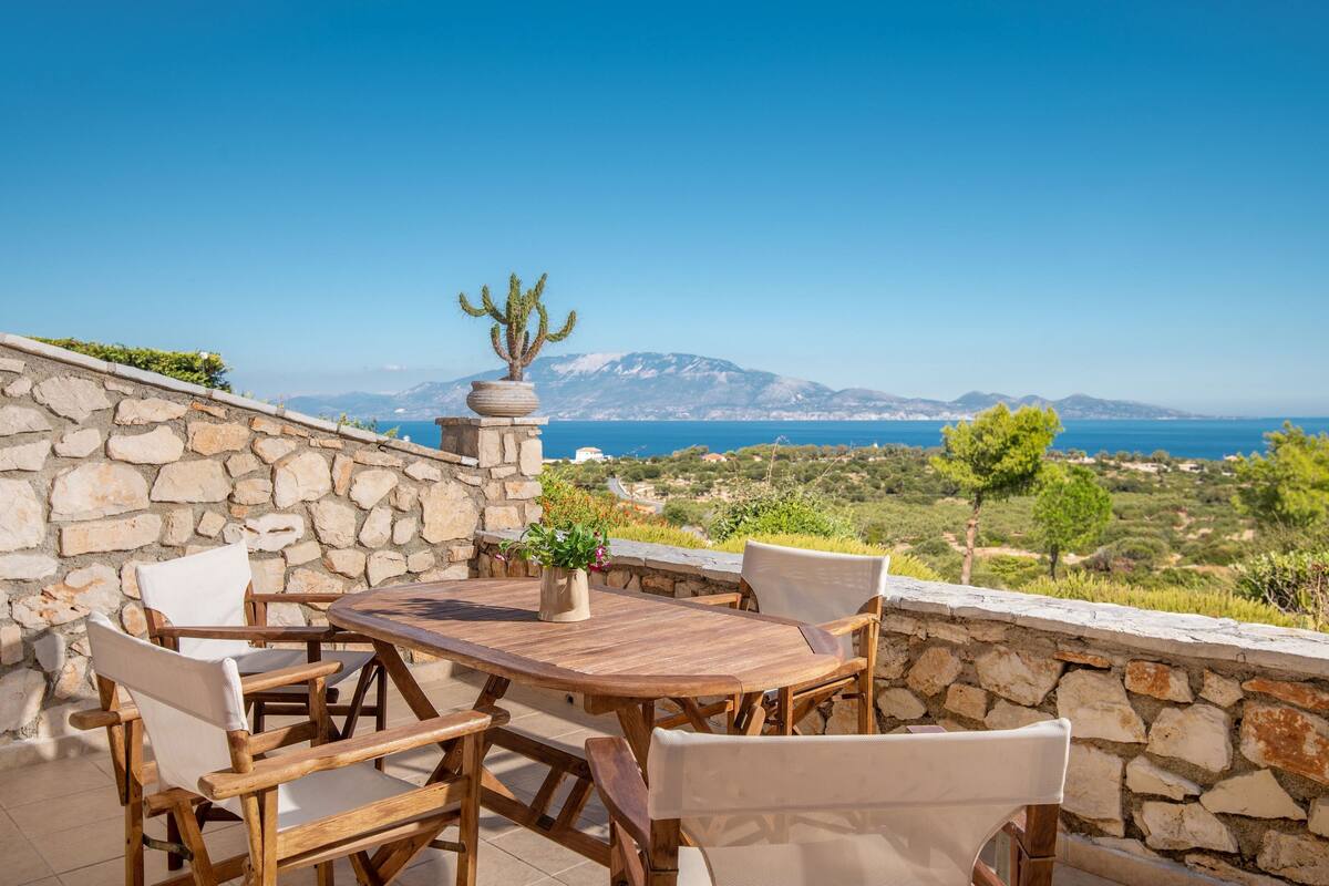 A wooden table and chairs set against a stone wall with a view of the sea and mountains.