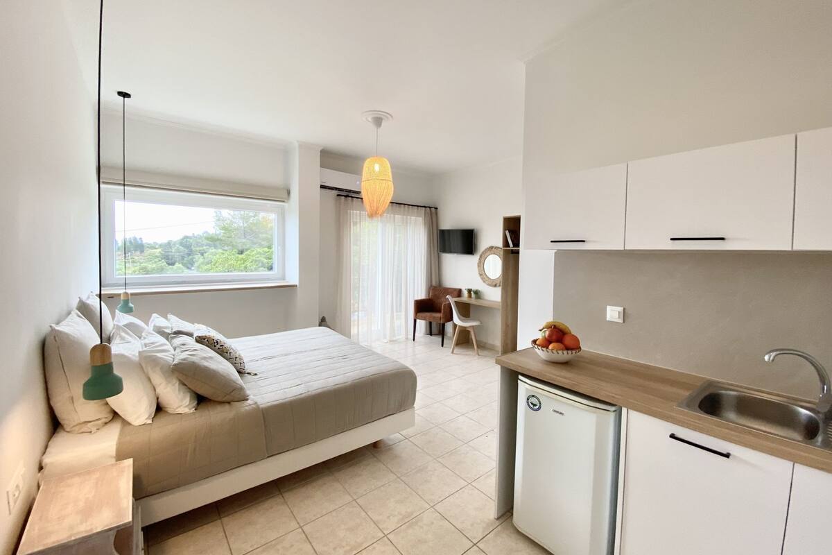 A modern bedroom with a bed, bedside table, and a window. Adjacent is a kitchenette with a sink, stove, and a small refrigerator.