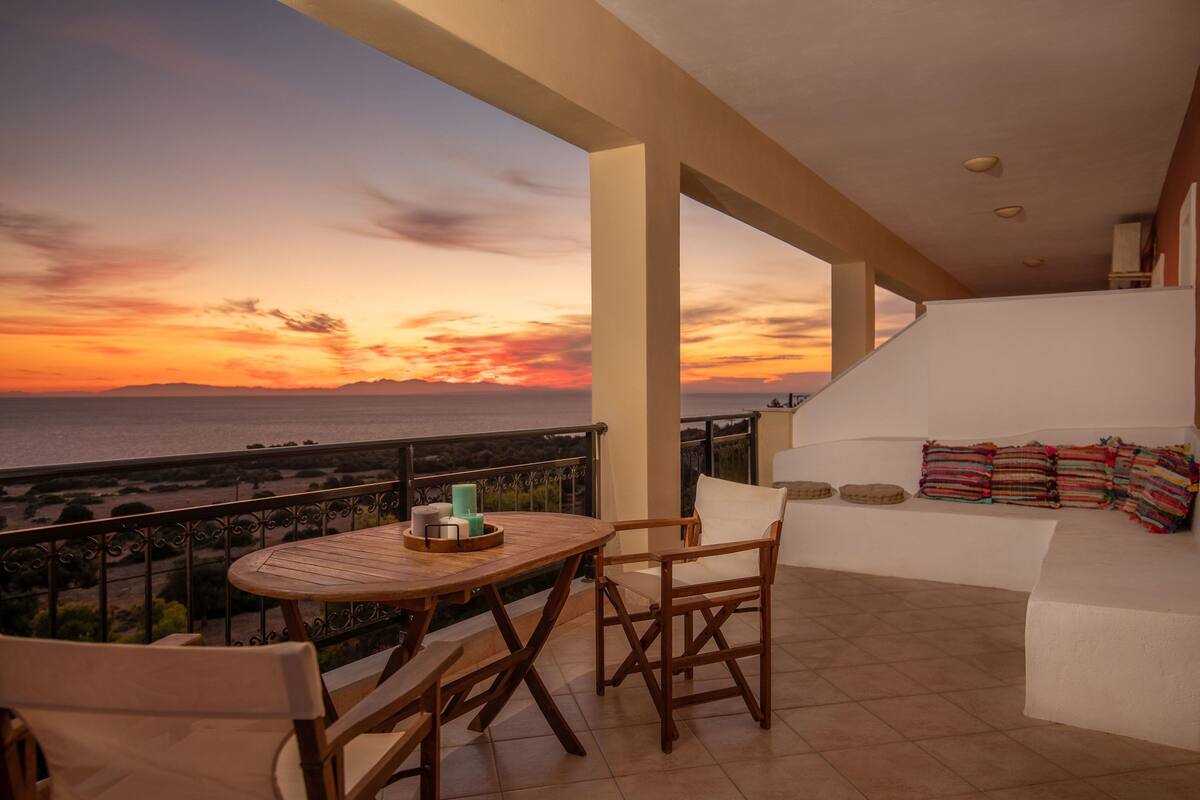 A balcony with a wooden table and chairs, a white sofa with colorful cushions, and a view of the sunset over the sea.