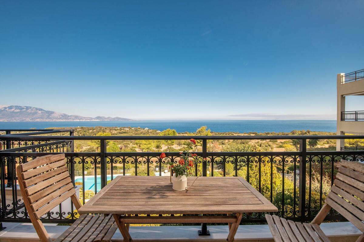 A wooden table with a vase of flowers on a balcony overlooking a pool and a scenic view of the sea and mountains.