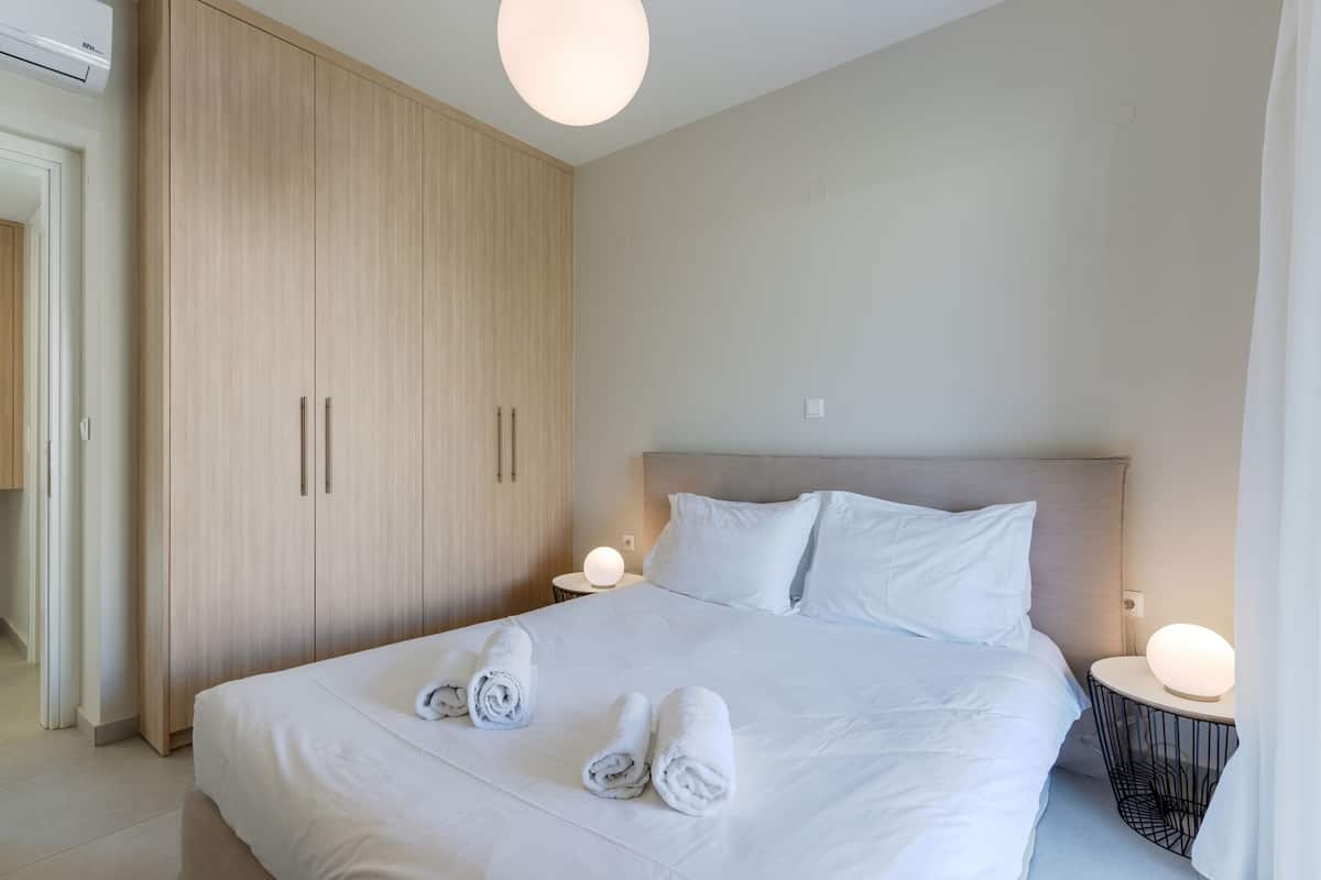 Una habitación de hotel moderna con una cama grande, dos mesitas de noche con lámparas, un armario y una paleta de colores neutros.