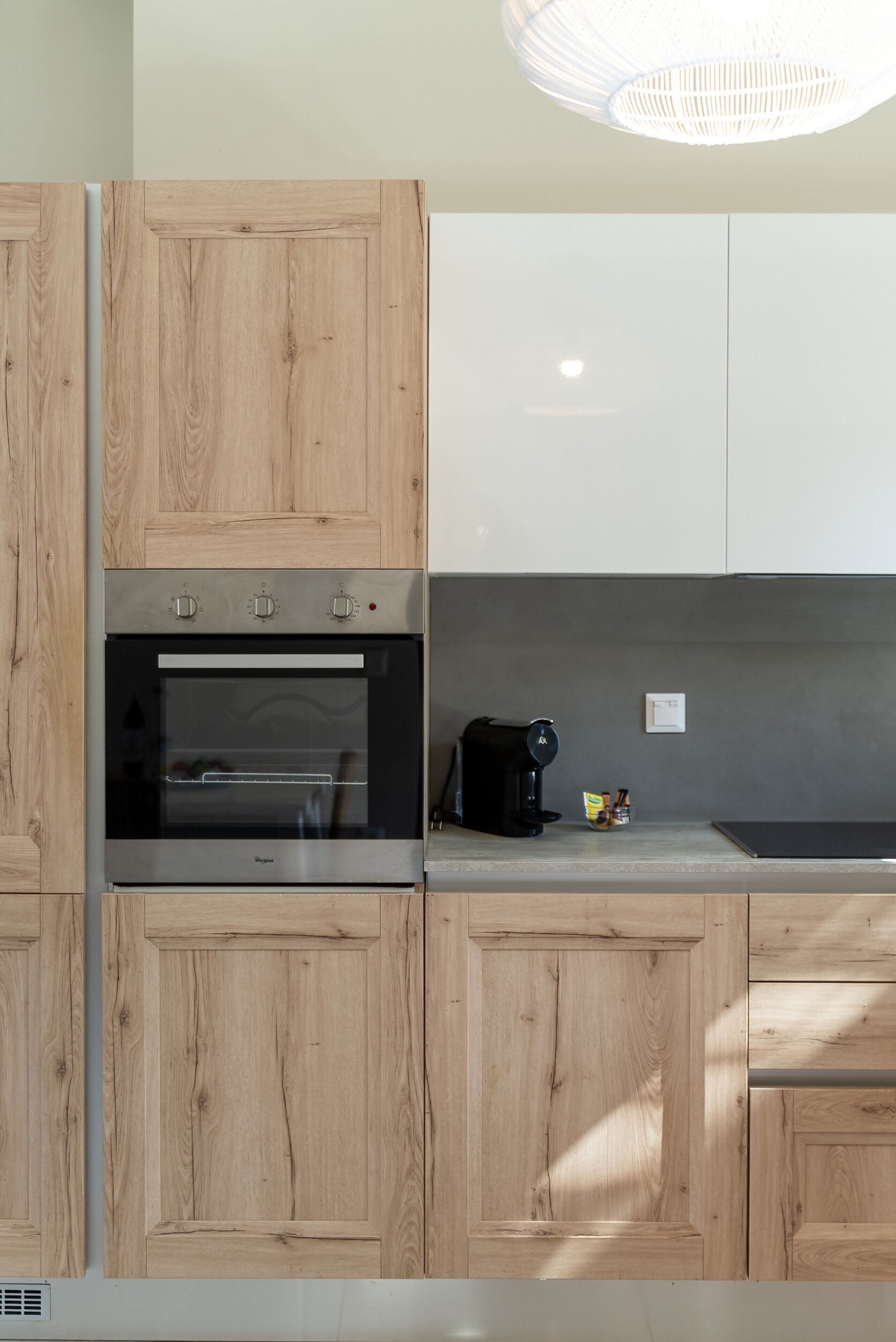 A modern kitchen with wooden cabinets, a built-in oven, a coffee maker, and a light fixture.
