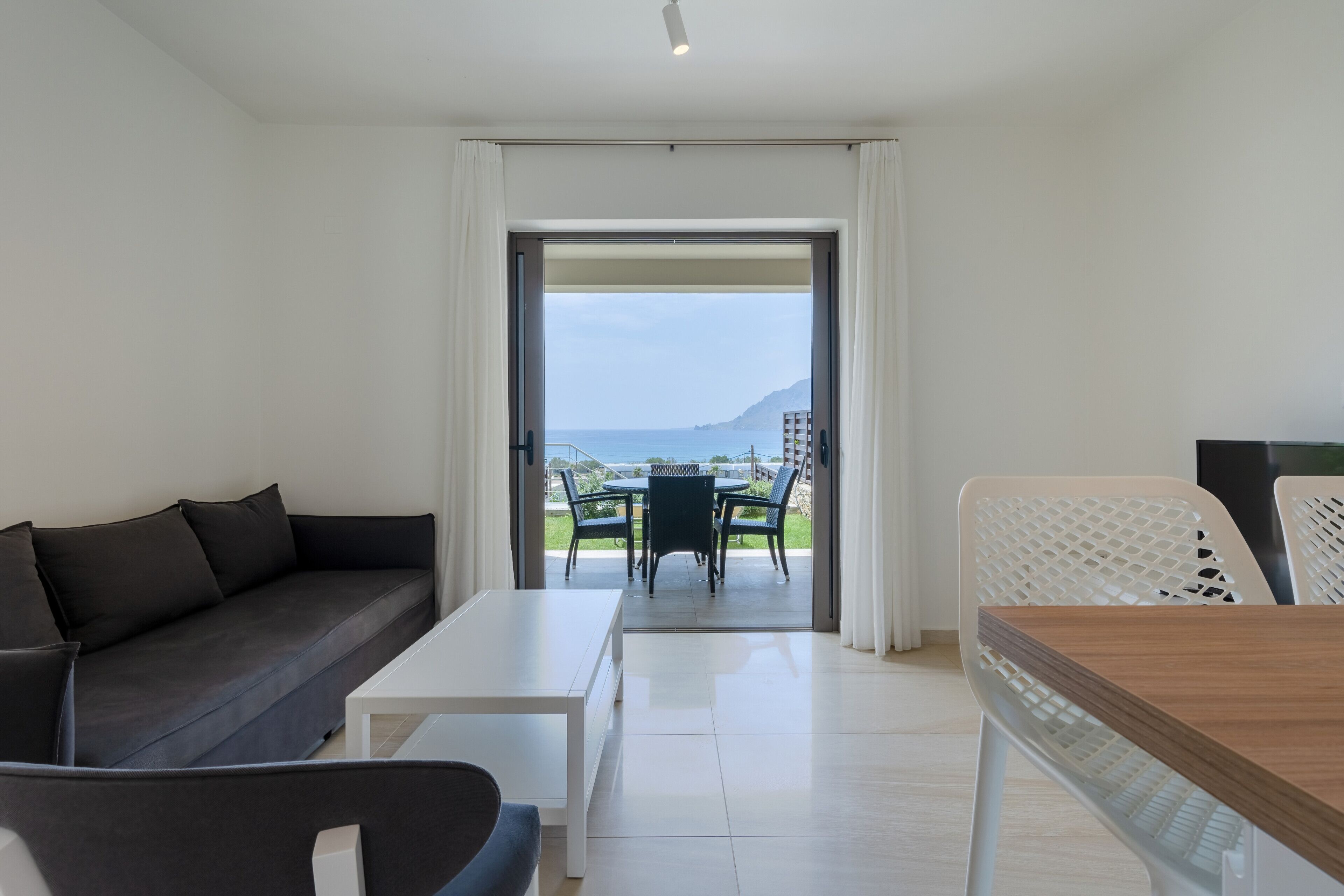 A modern living room with a sofa, a coffee table, and a dining area with a table and chairs. A large window provides a view of the outdoors.