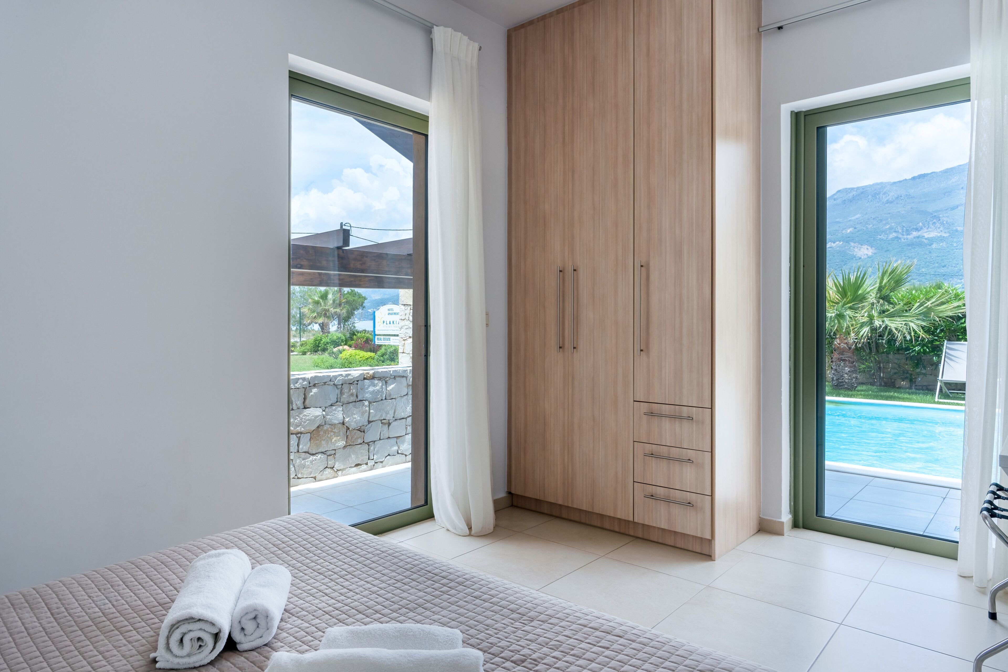 A bedroom with a bed, a wooden wardrobe, and a view of a swimming pool.