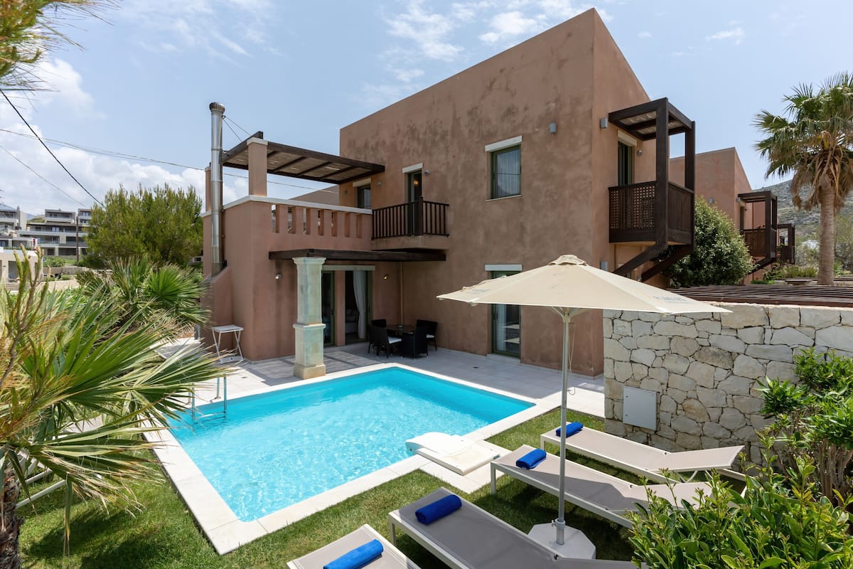 Una casa moderna de dos pisos con piscina, mobiliario exterior y un muro de piedra.