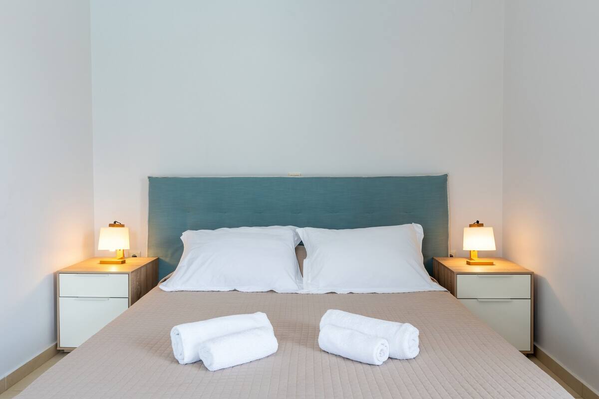 Una cama cuidadosamente tendida con dos almohadas y dos toallas enrolladas, flanqueada por mesitas de noche con lámparas.