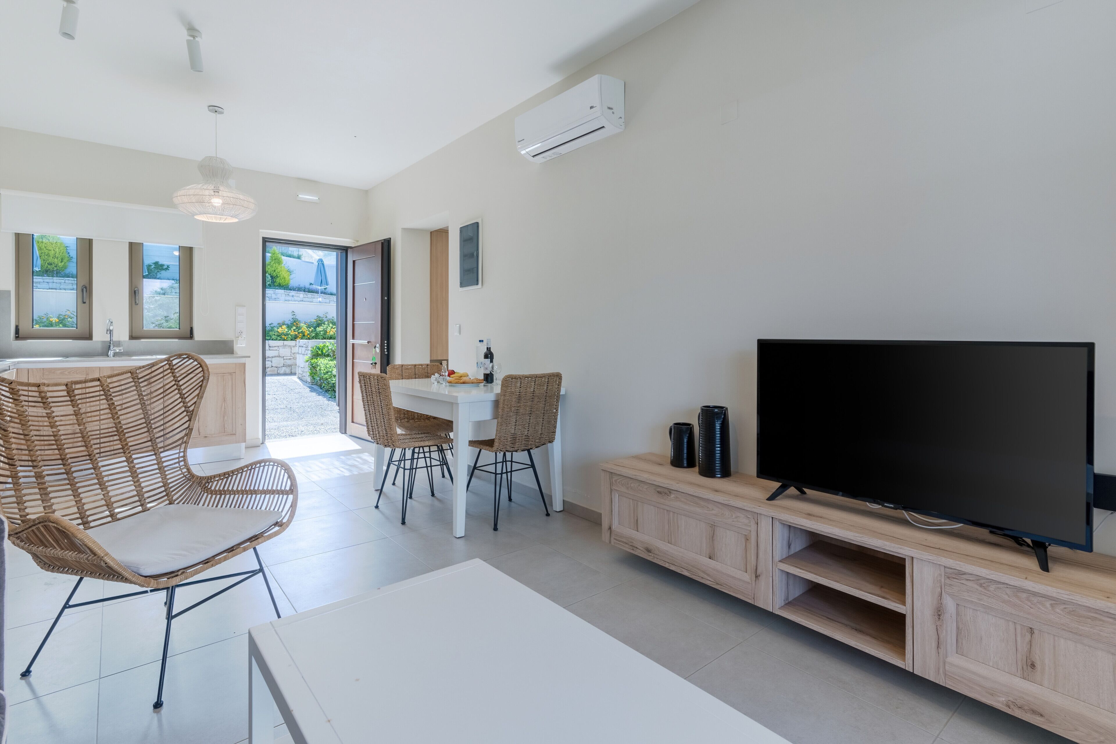 A modern living room with a large flat-screen TV, wicker chairs, a dining table, and a kitchen area with a view of the outdoors.
