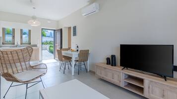 A modern living room with a large flat-screen TV, wicker chairs, a dining table, and a kitchen area with a view of the outdoors.