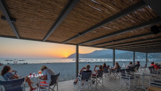 Una terraza de madera con mesas y sillas da a una playa al atardecer.