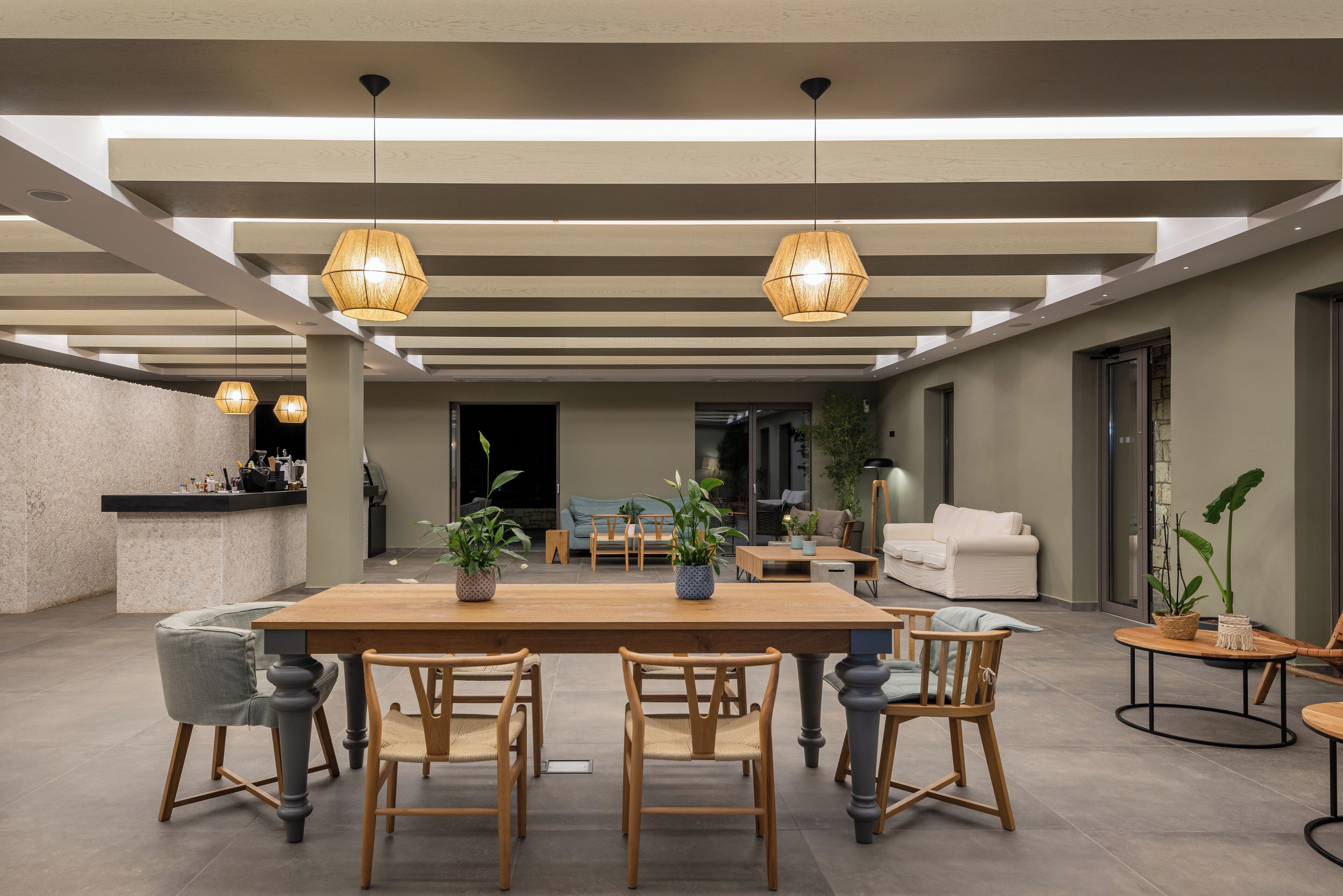 A spacious living area with a wooden dining table, chairs, and hanging pendant lights. There is a sofa and a coffee table in the background.