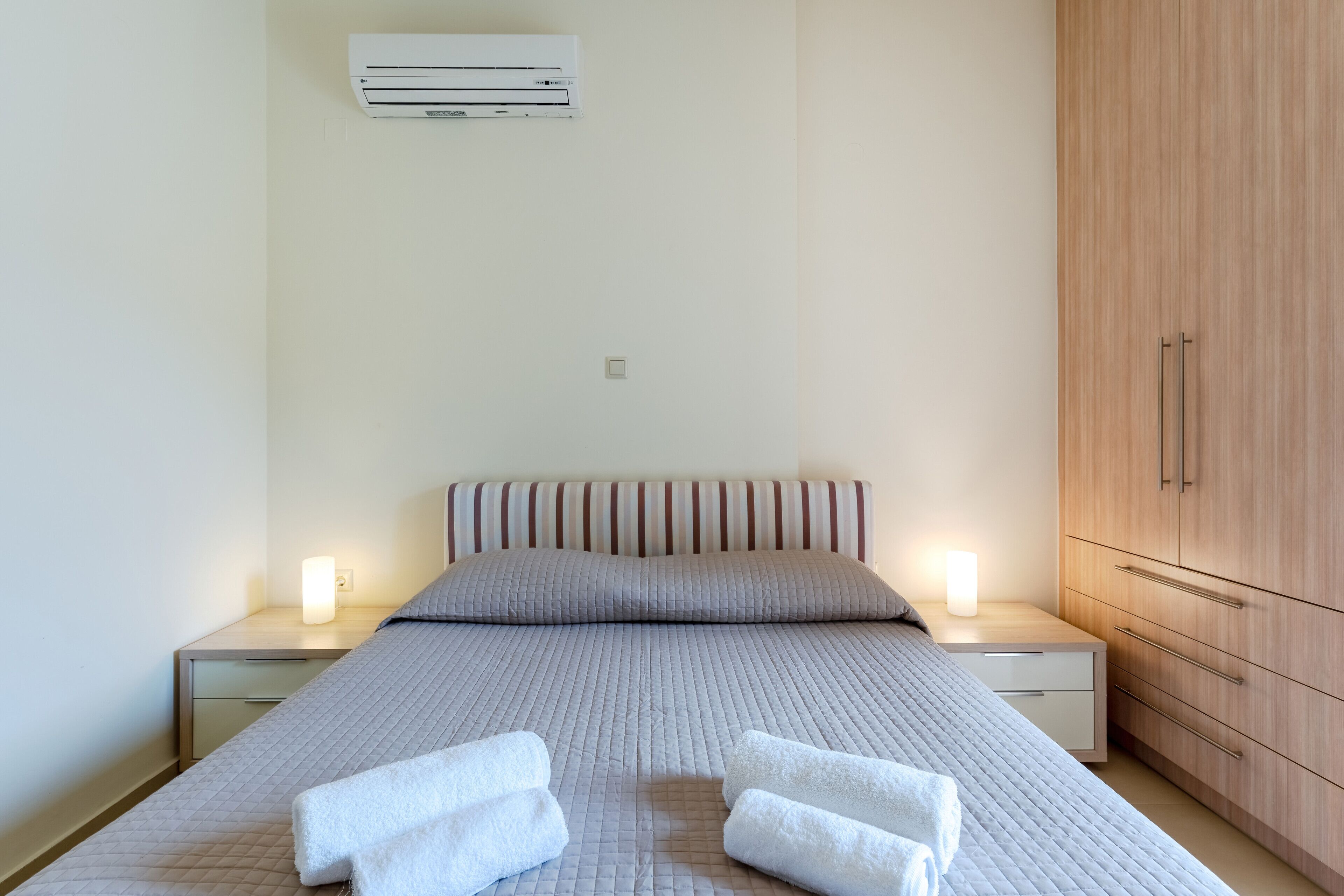 A neatly made bed with pillows and two white towels, a bedside table with a lamp, a wooden wardrobe, and an air conditioning unit on the wall.