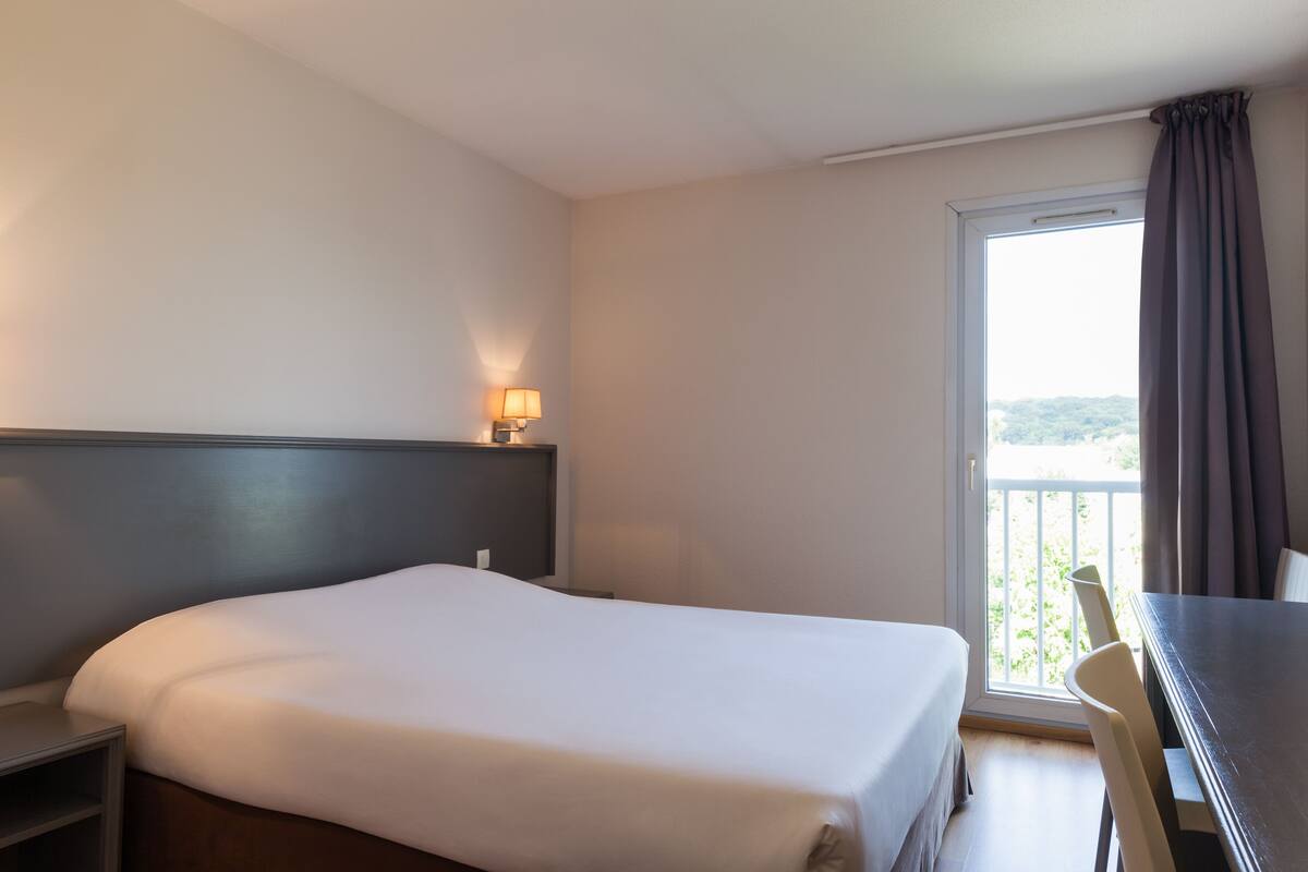 A hotel room with a bed, a desk, a lamp, and a window with curtains.