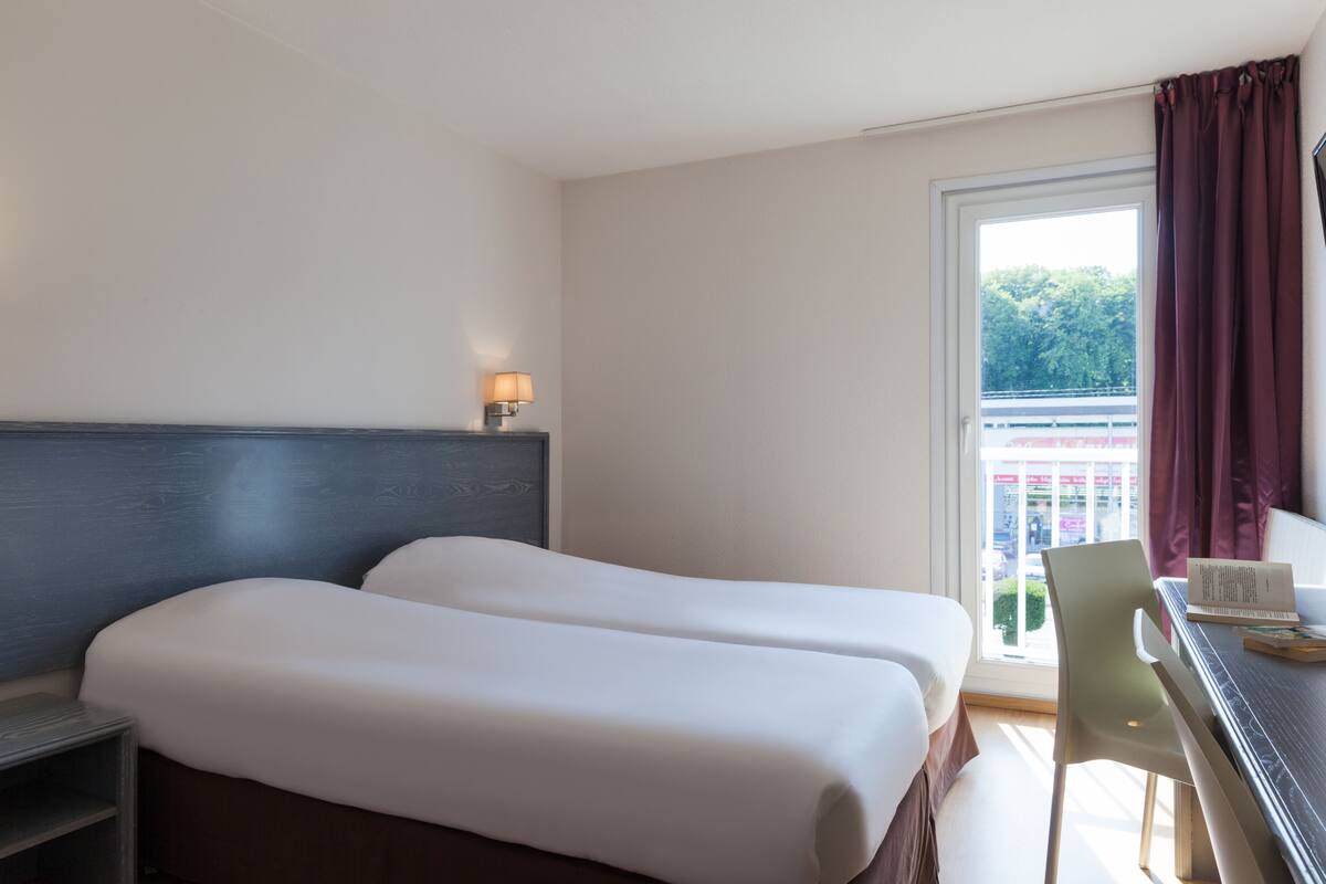 A hotel room with two beds, a desk, a chair, a lamp, and a window with curtains.