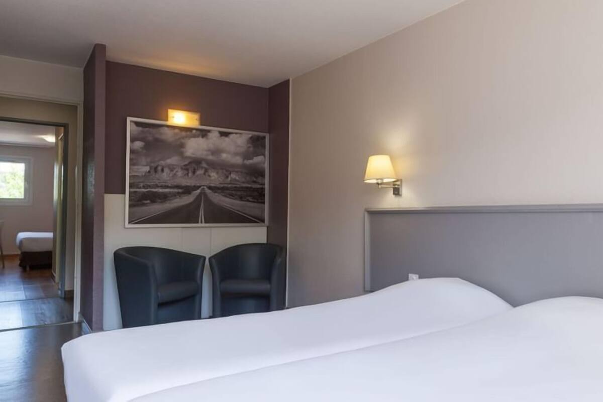 A hotel room with a large bed, two chairs, a wall-mounted lamp, and a framed picture of a road.