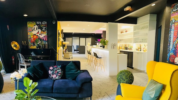 A modern living room with a dark blue sofa, a yellow armchair, and a glass coffee table with plants. A kitchen and dining area are visible in the background.