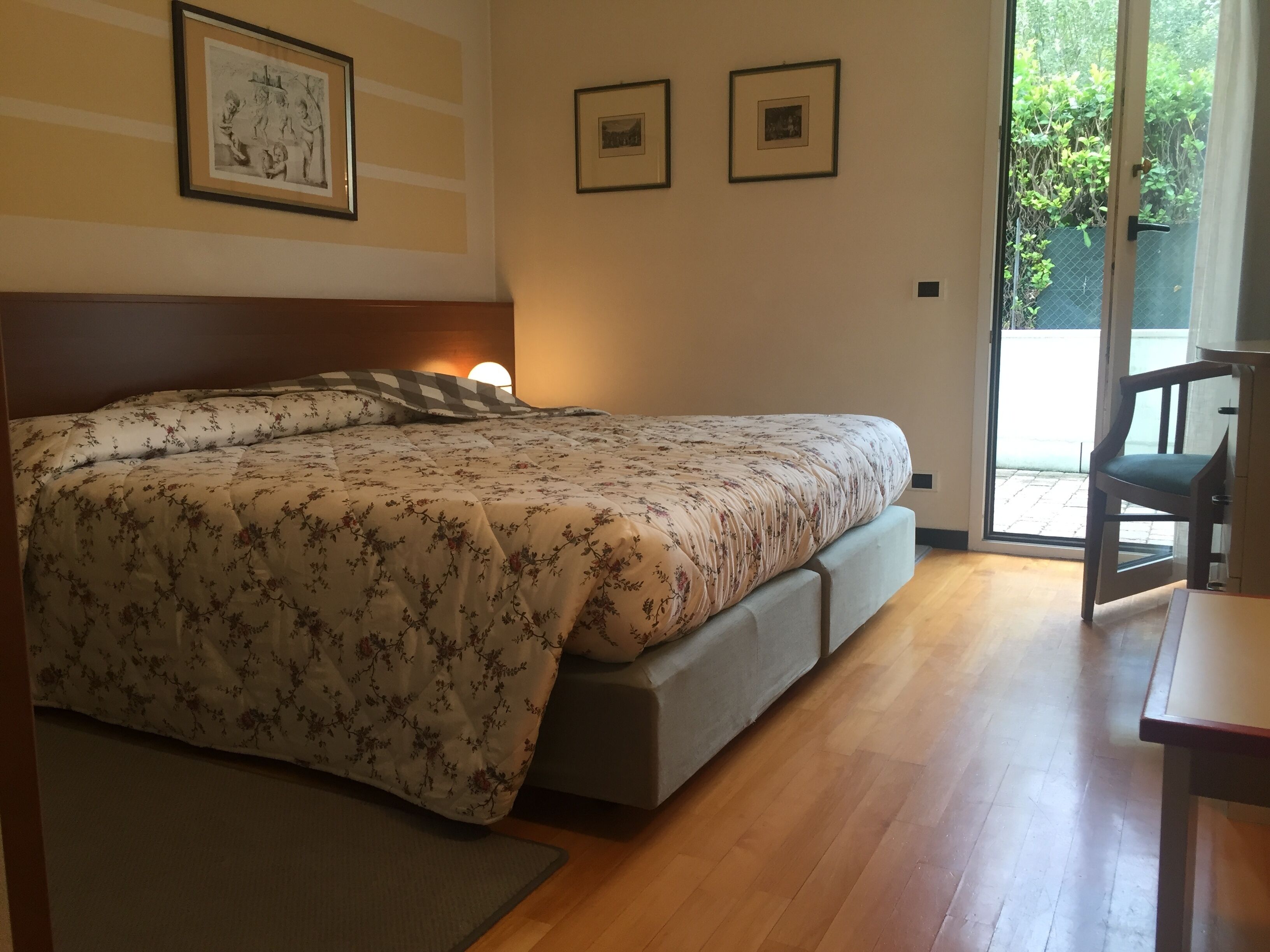 Quarto com cama, cadeira e mesa. Há quadros nas paredes e uma janela com vista para o verde.