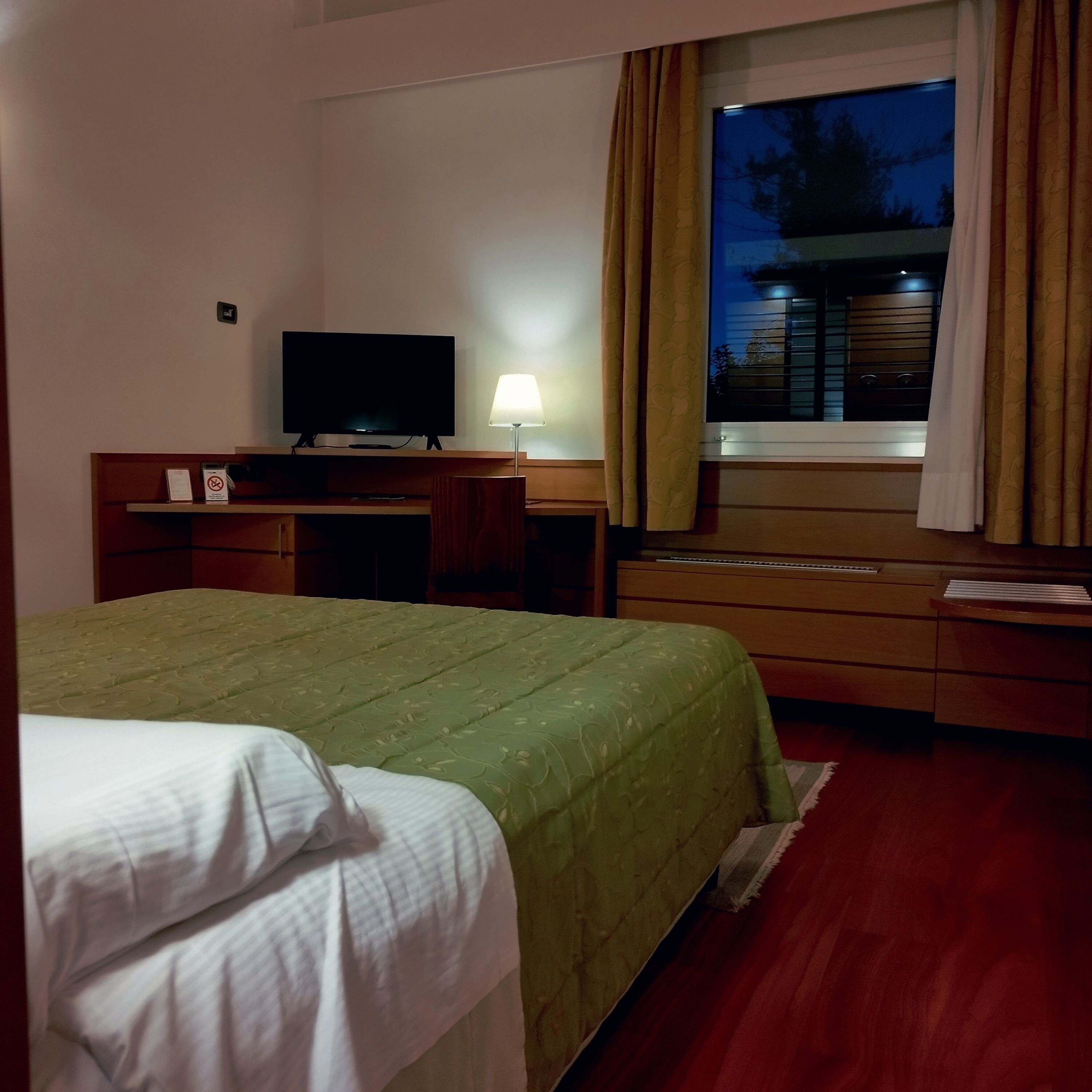 Une chambre d’hôtel avec un lit, un bureau doté d’une télévision, une lampe et une fenêtre avec des rideaux.