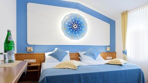 Habitación de hotel con un reloj de pared grande, cama con ropa de cama azul, cabecera de madera, mesitas de noche y una ventana con cortinas.