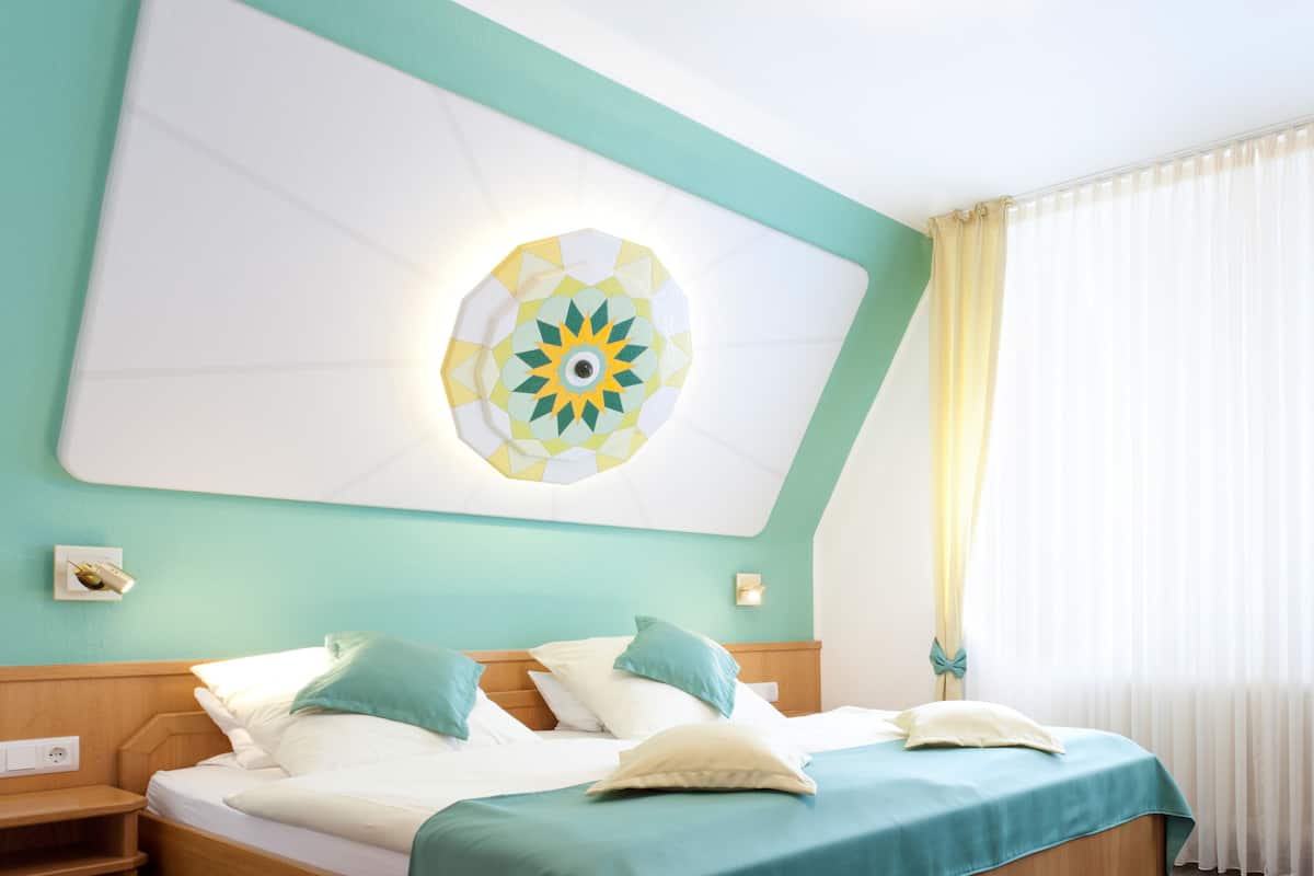 Cama bem arrumada com colcha verde-água e várias almofadas, um objeto decorativo fixado na parede e uma janela com cortinas finas.