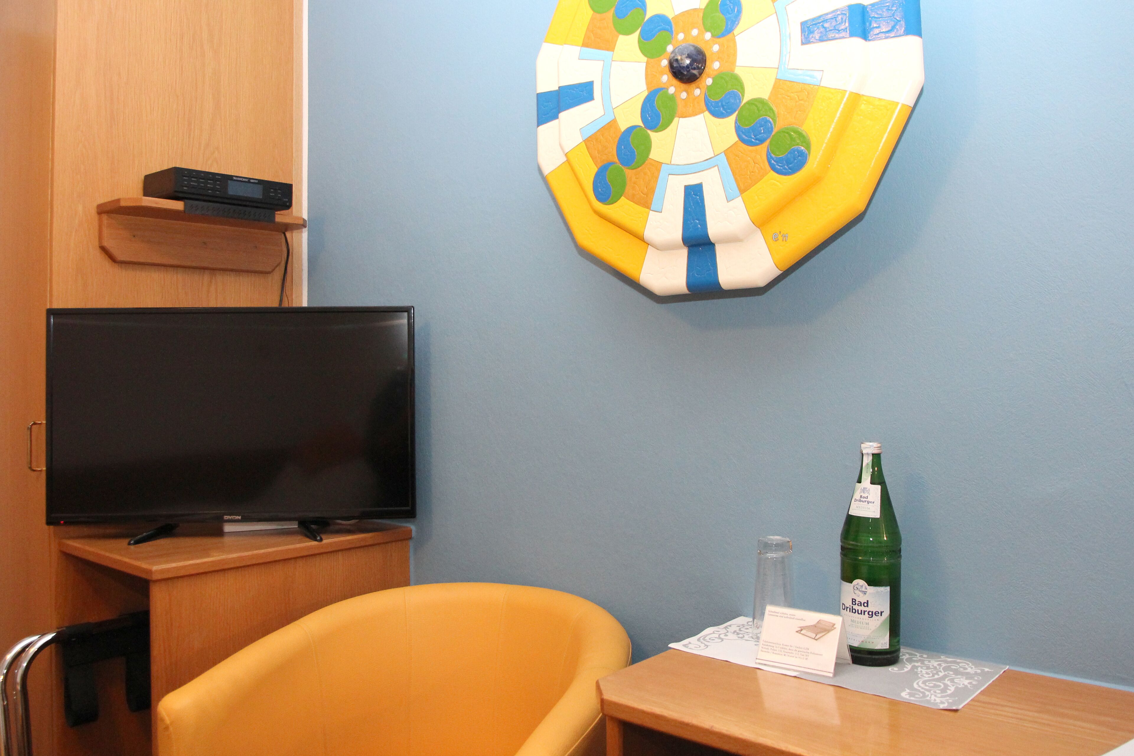 A hotel room with a television on a wooden stand, a yellow armchair, a small table with a bottle and a coaster, and a wall decoration.
