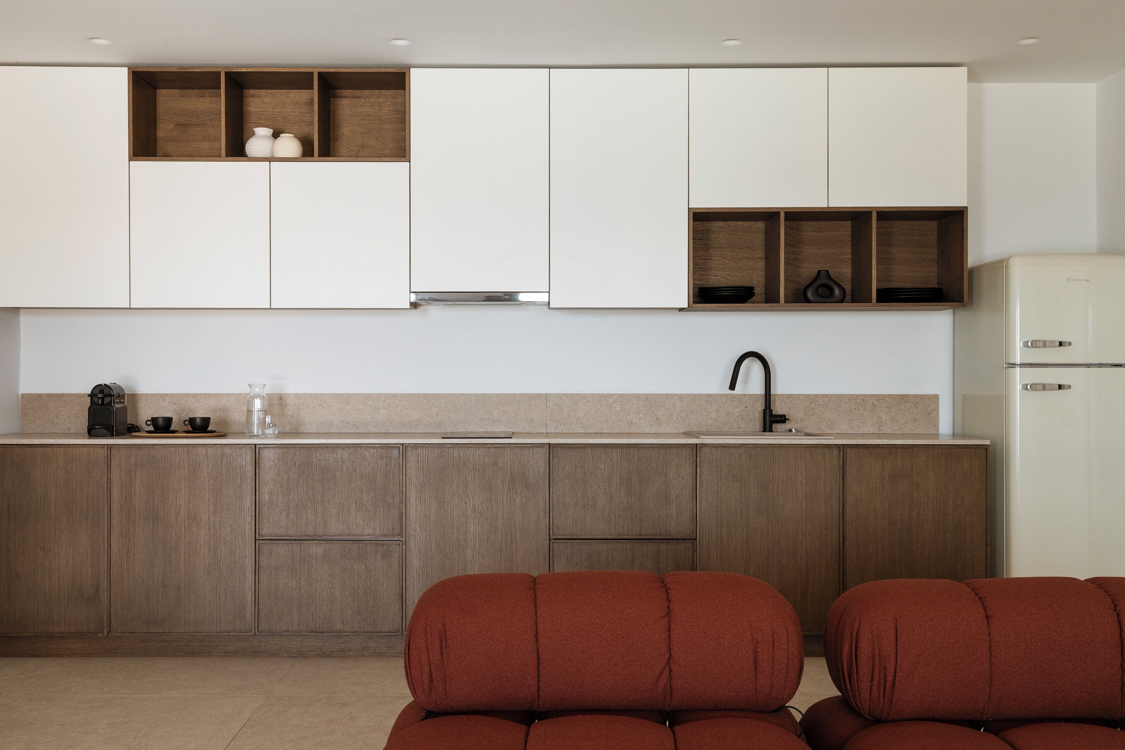 Cocina moderna con armarios de madera, encimera blanca y horno empotrado. Hay dos sillas rojas en primer plano.