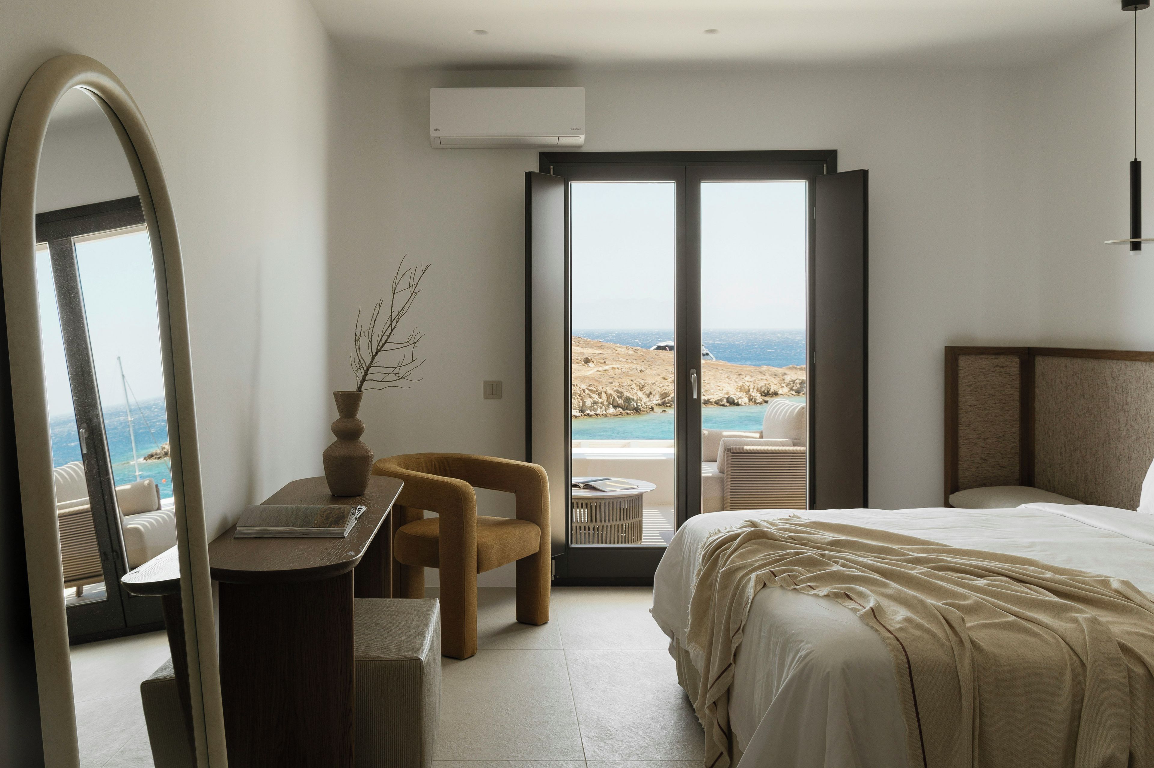A hotel room with a bed, a desk, a chair, a large mirror, and a view of the sea.