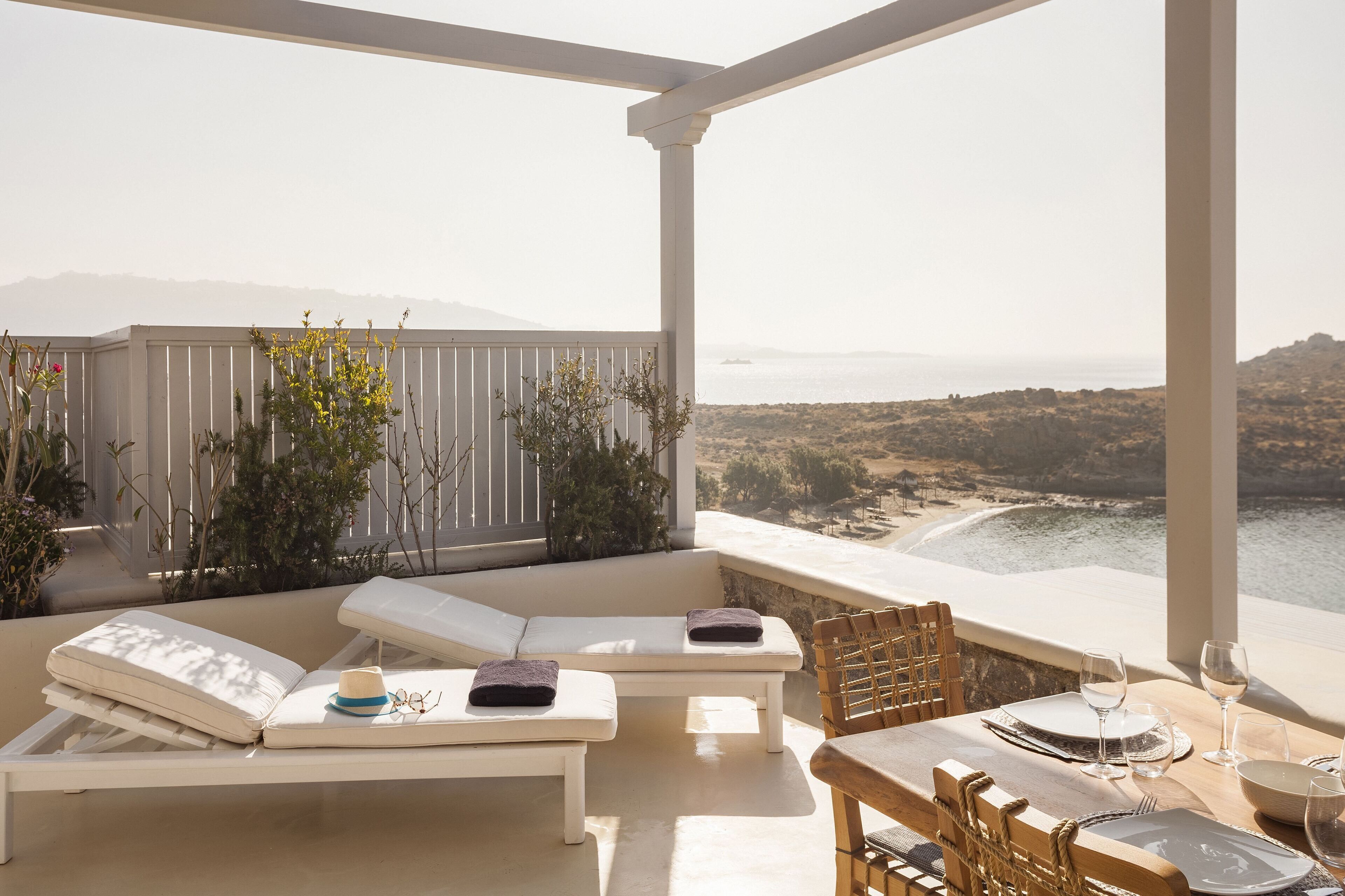 A sunlit balcony with white cushioned lounge chairs, a dining table set for two, and a view of the sea and hills.