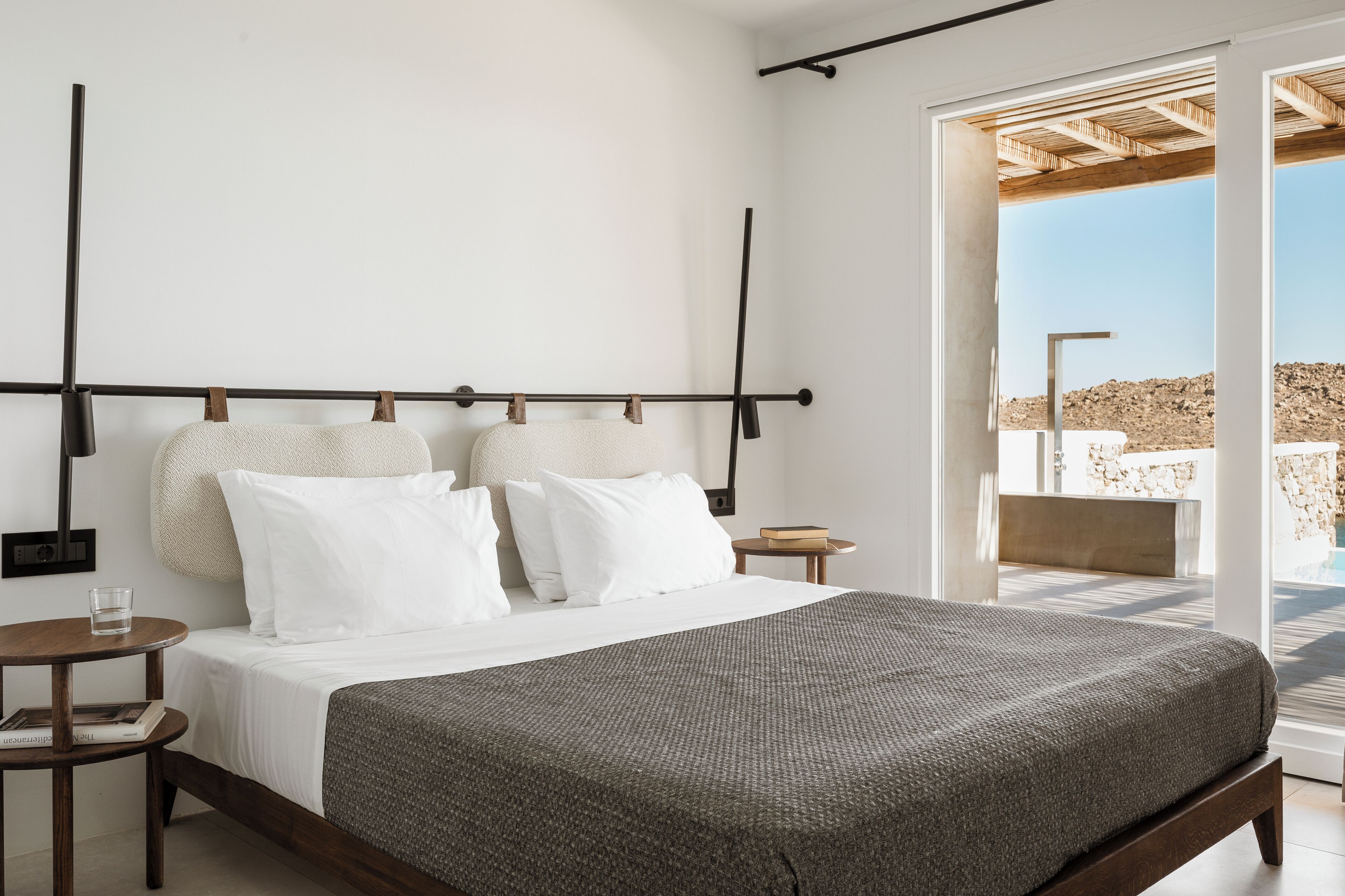 Une chambre moderne avec un grand lit, une tête de lit en bois, une table de chevet avec un verre d’eau, et une vue sur l’extérieur grâce à une porte vitrée.