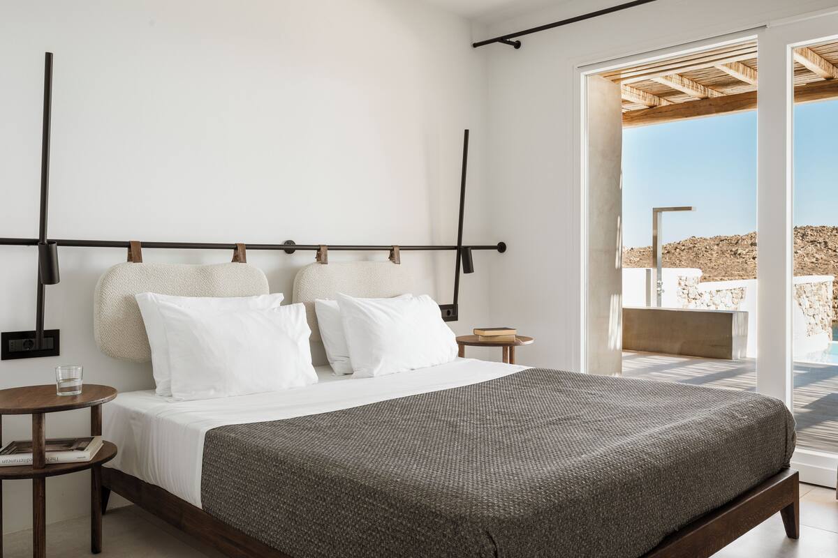 Une chambre moderne avec un grand lit, une tête de lit en bois, une table de chevet avec un verre d’eau, et une vue sur l’extérieur grâce à une porte vitrée.