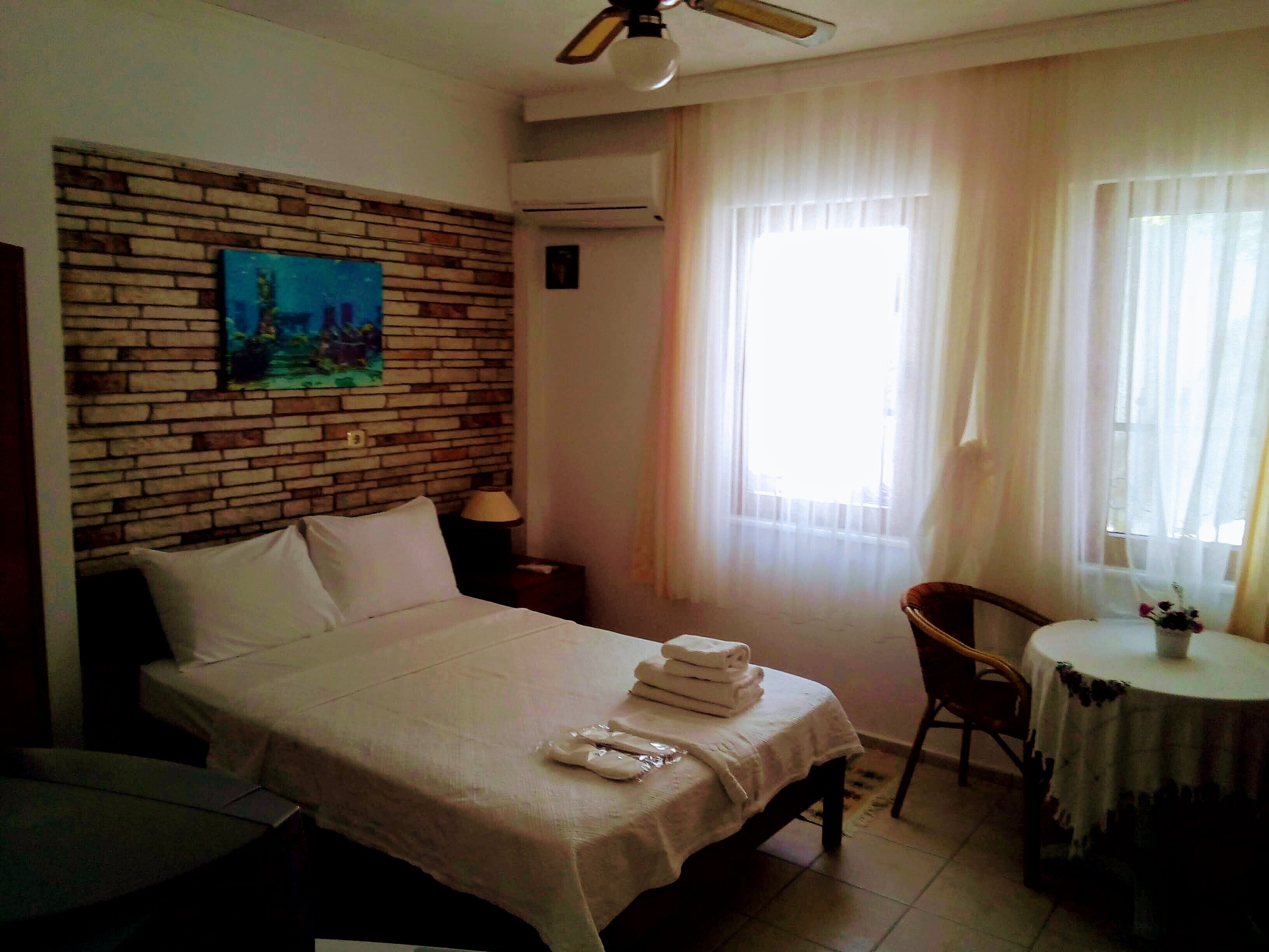 Habitación de hotel con una cama, una silla, una mesa con un jarrón, un cuadro en la pared y una ventana con cortinas.