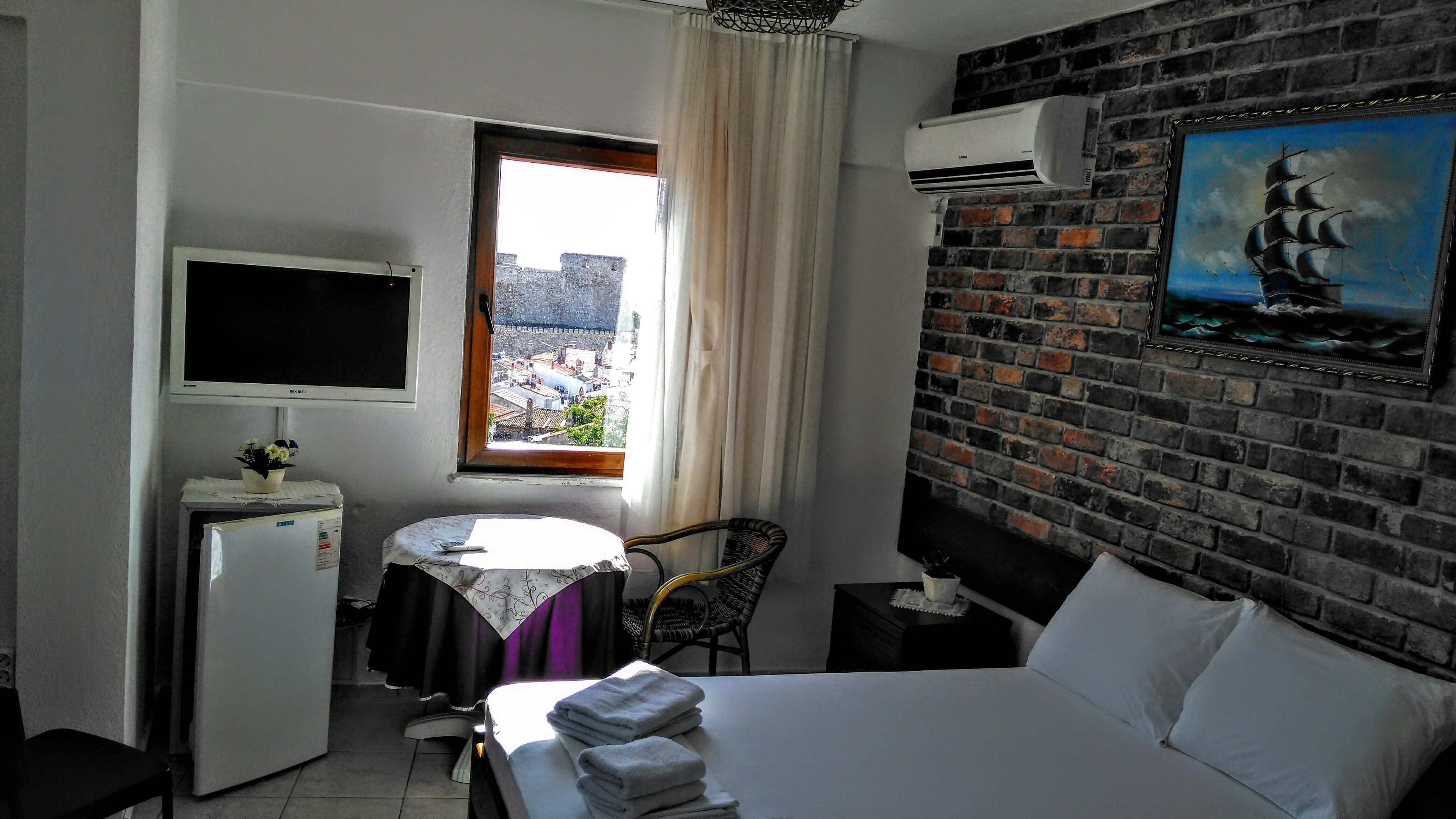 A hotel room with a bed, a desk, a chair, a TV, an air conditioner, and a window with a view of buildings.
