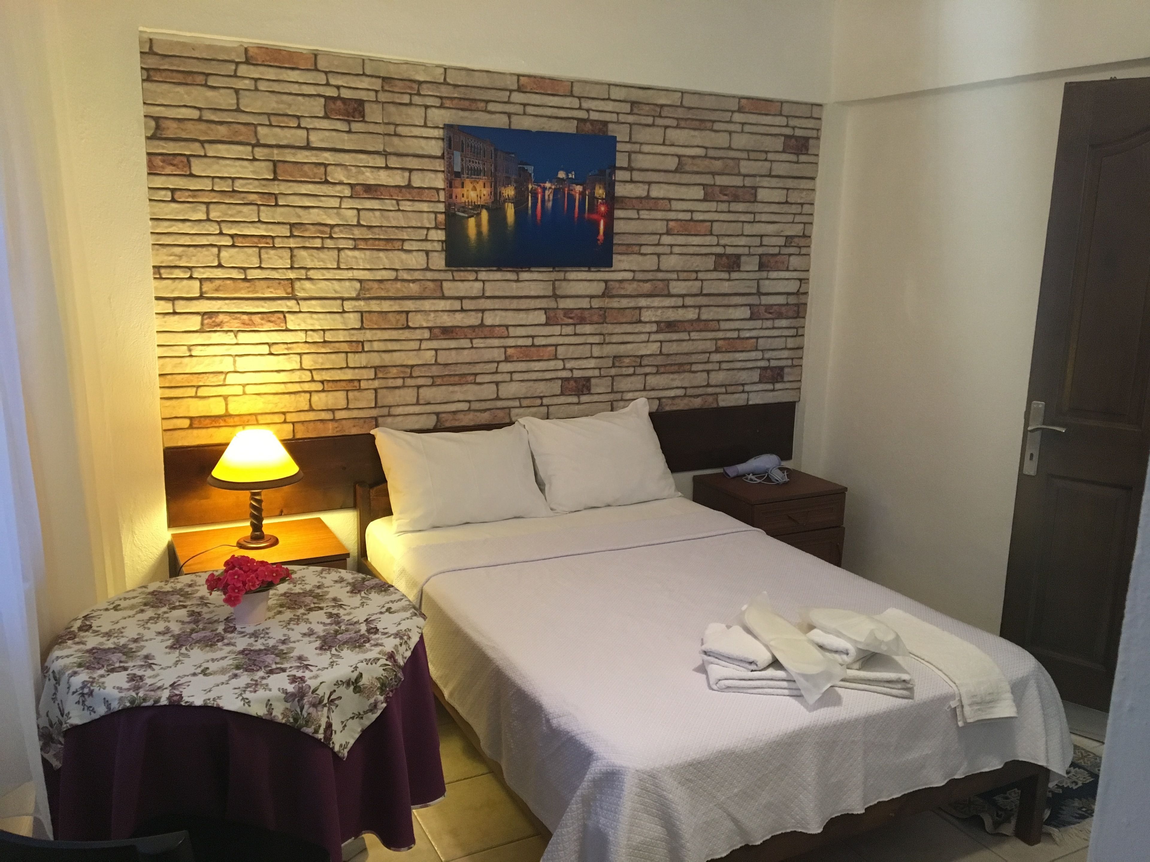 A hotel room with a brick wall, a bed with white linens, a nightstand with a lamp, and a painting of a cityscape.