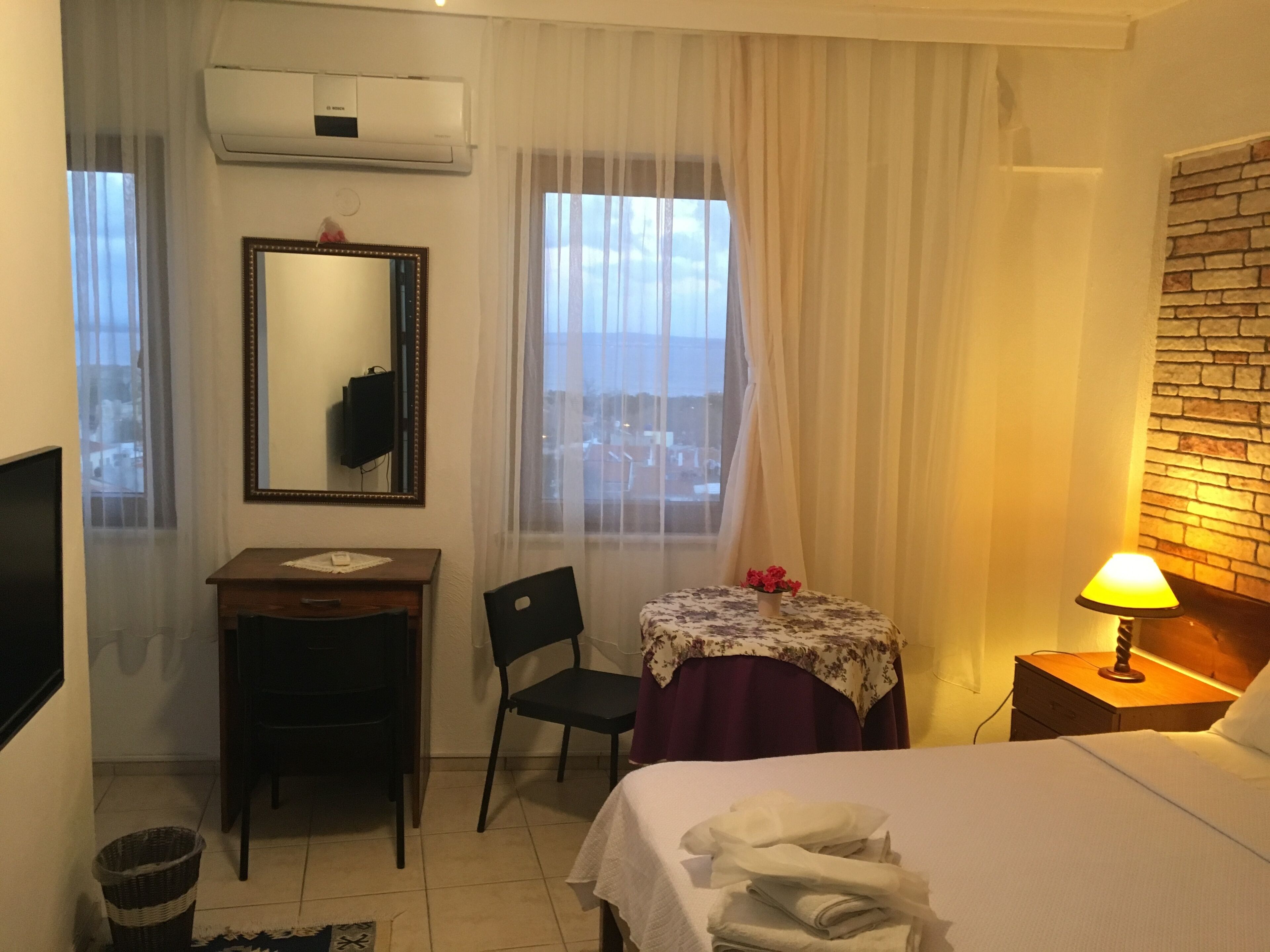 A hotel room with a bed, a desk with a chair, a small table with a lamp, and a window with curtains.