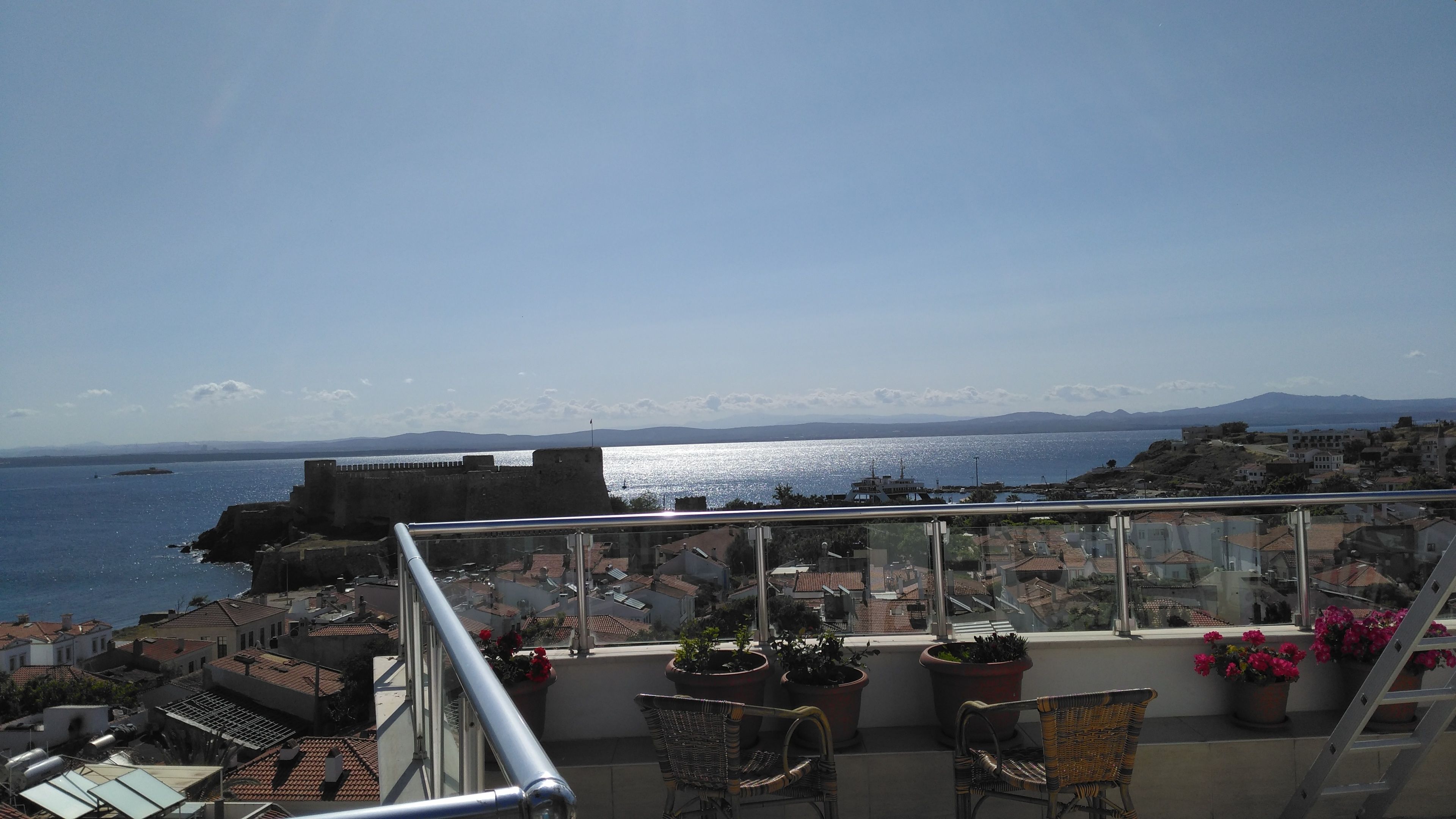 A balcony with a view of the sea and distant mountains. There are potted plants and wicker chairs on the balcony.